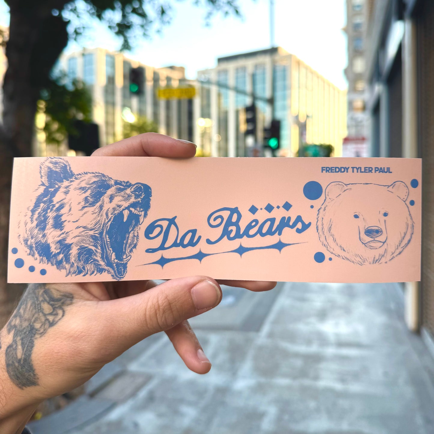 Hand holding a pink card with blue bear illustrations and 'Da Bears' text against an urban background.