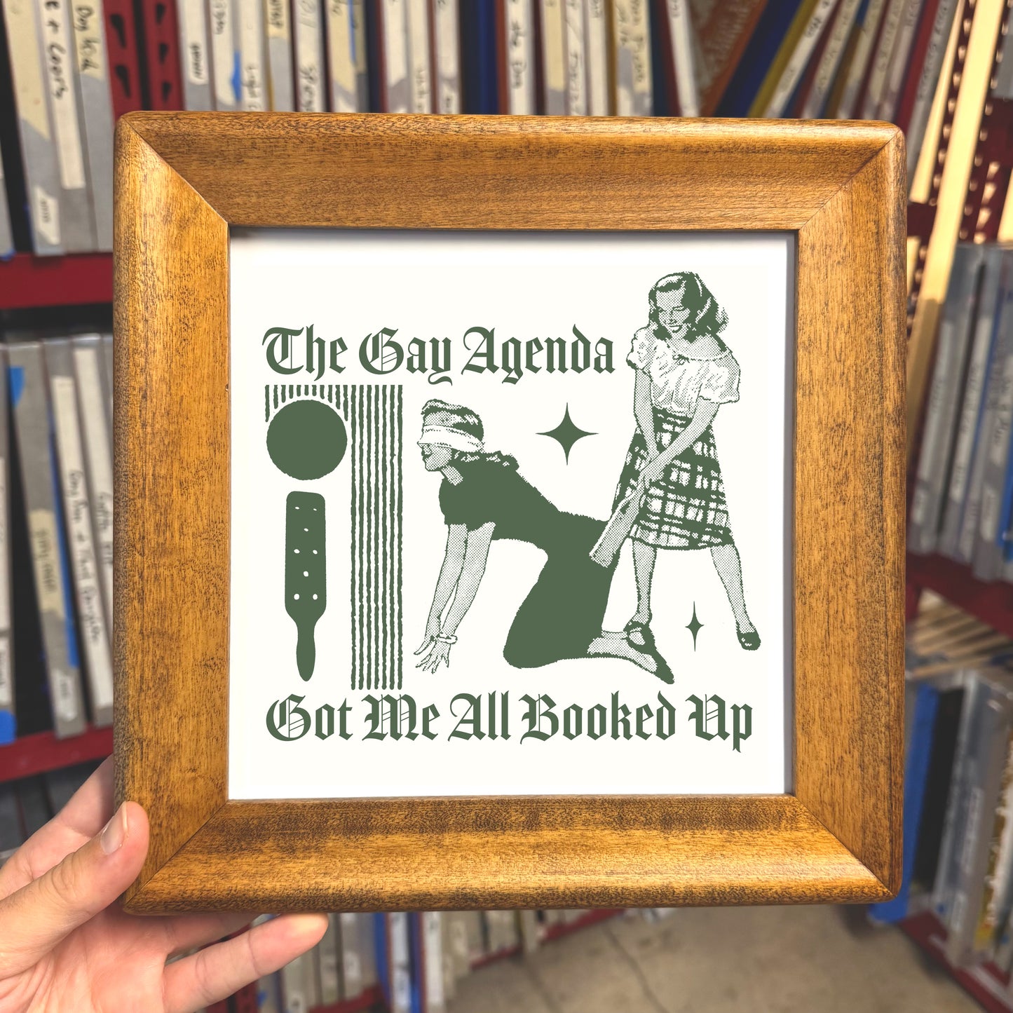 Framed poster with 'The Gay Agenda' text and illustrations held in front of a bookshelf.