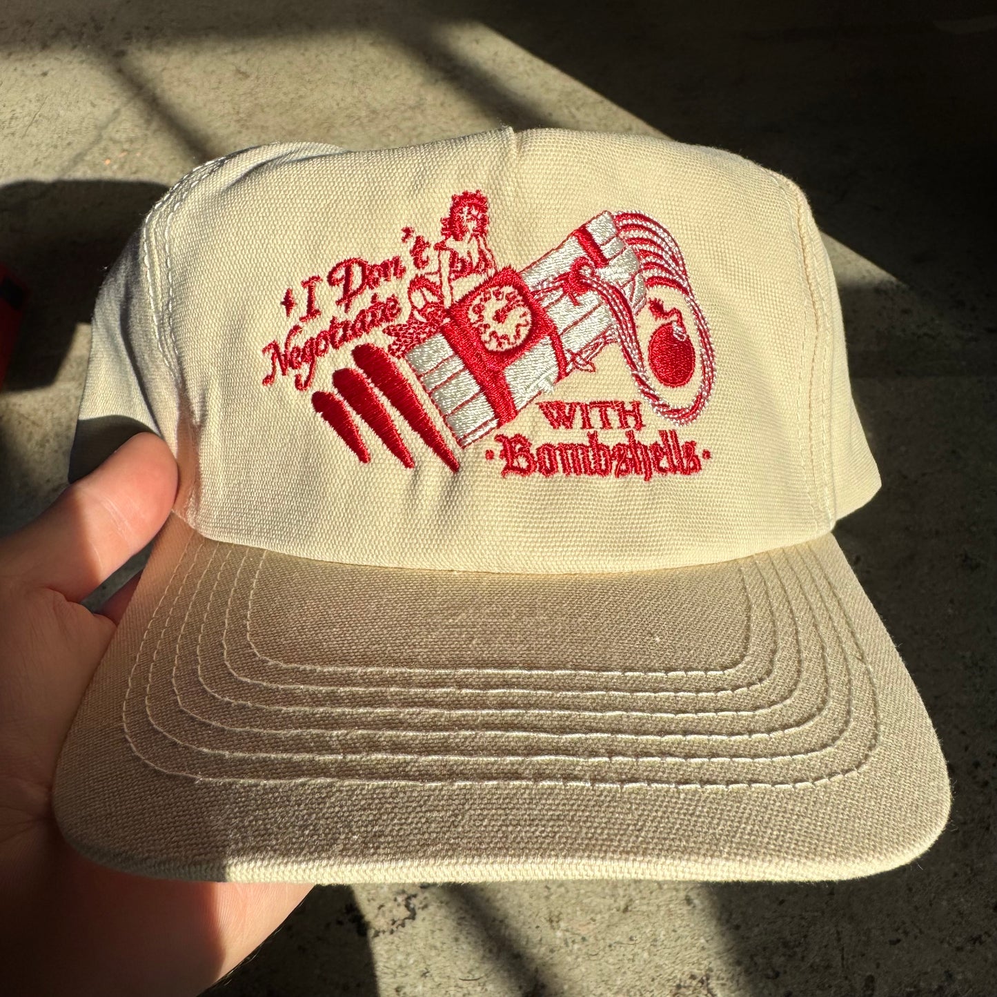 Beige cap with red embroidered text and design held by a hand on a concrete surface.