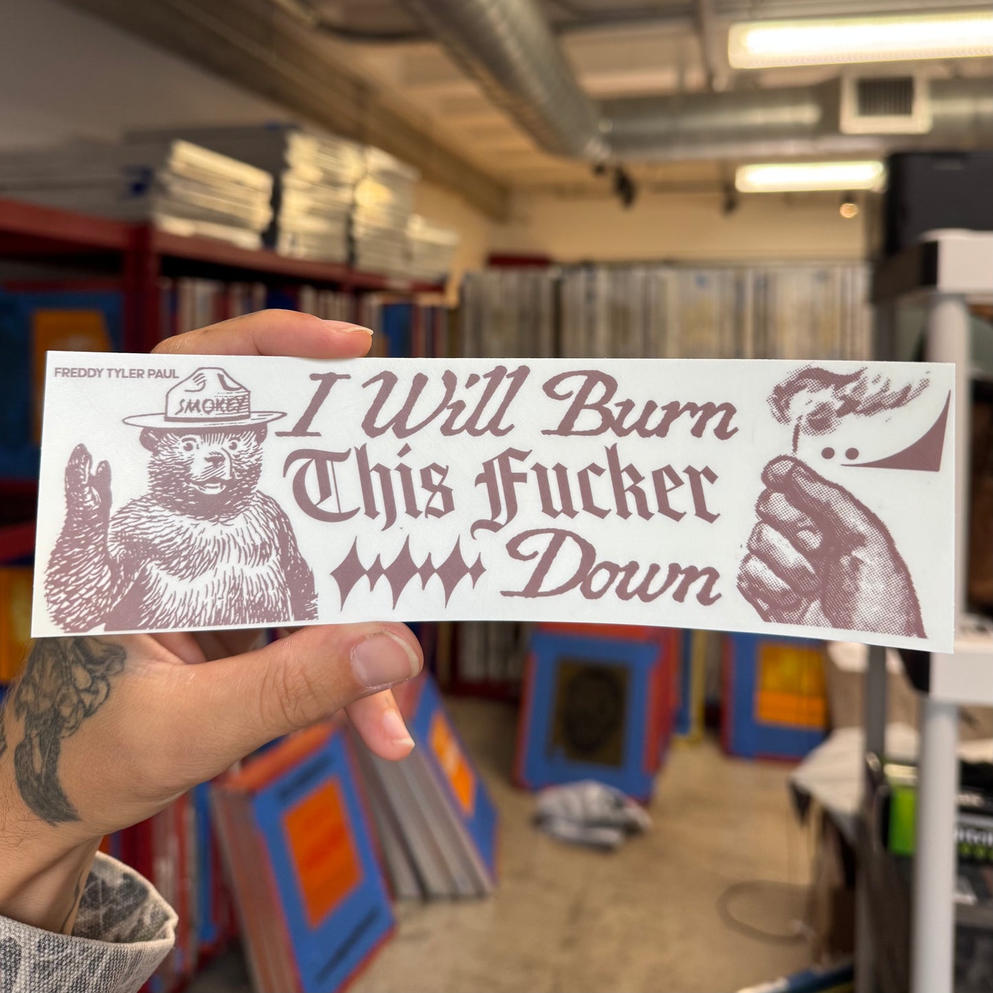 Hand holding a sticker with graphic text and bear illustration in an indoor setting.