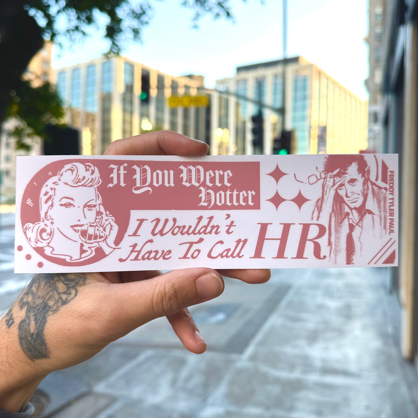 Hand holding a humorous sticker with text and graphics in an urban setting