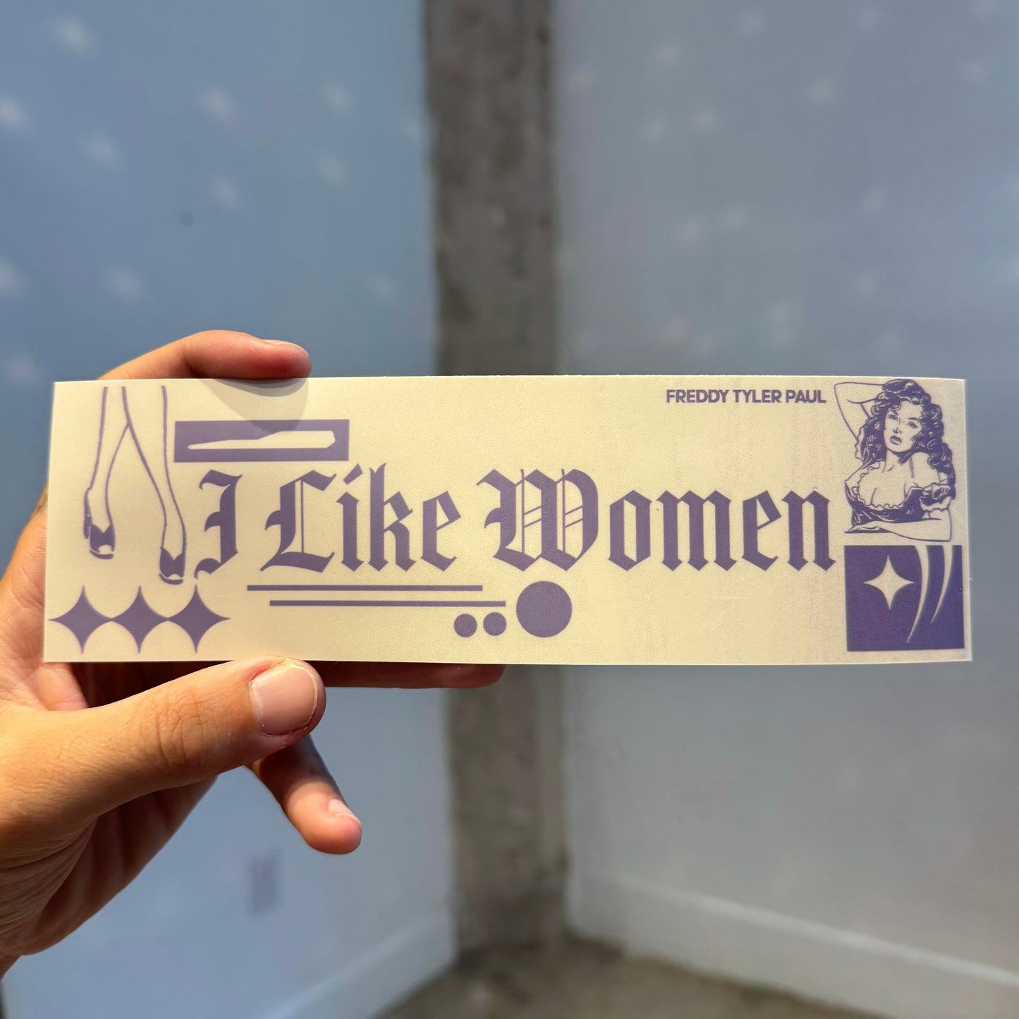 Hand holding a bumper sticker with text and graphics against a light blue wall.