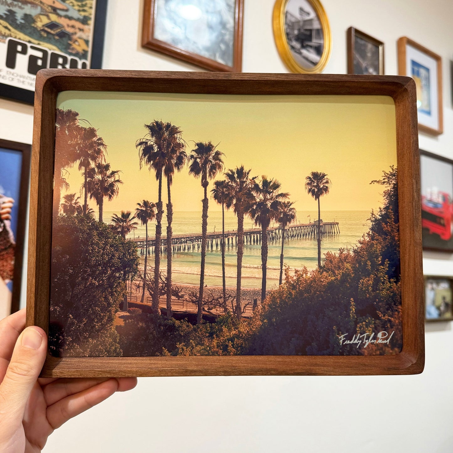Wooden framed artwork of a beach scene with palm trees and a pier.