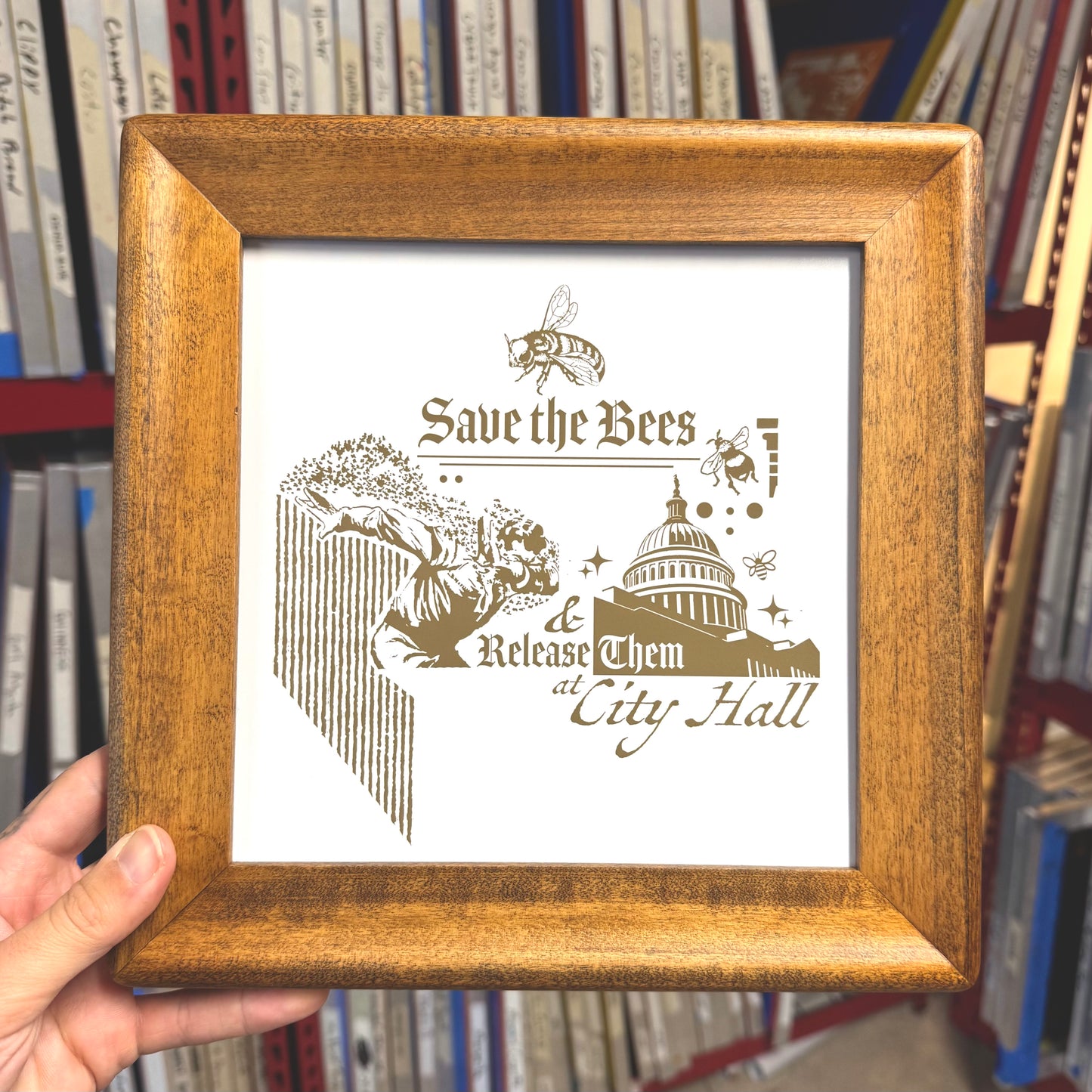 Wooden framed print with 'Save the Bees' text and graphic, held in front of bookshelf.