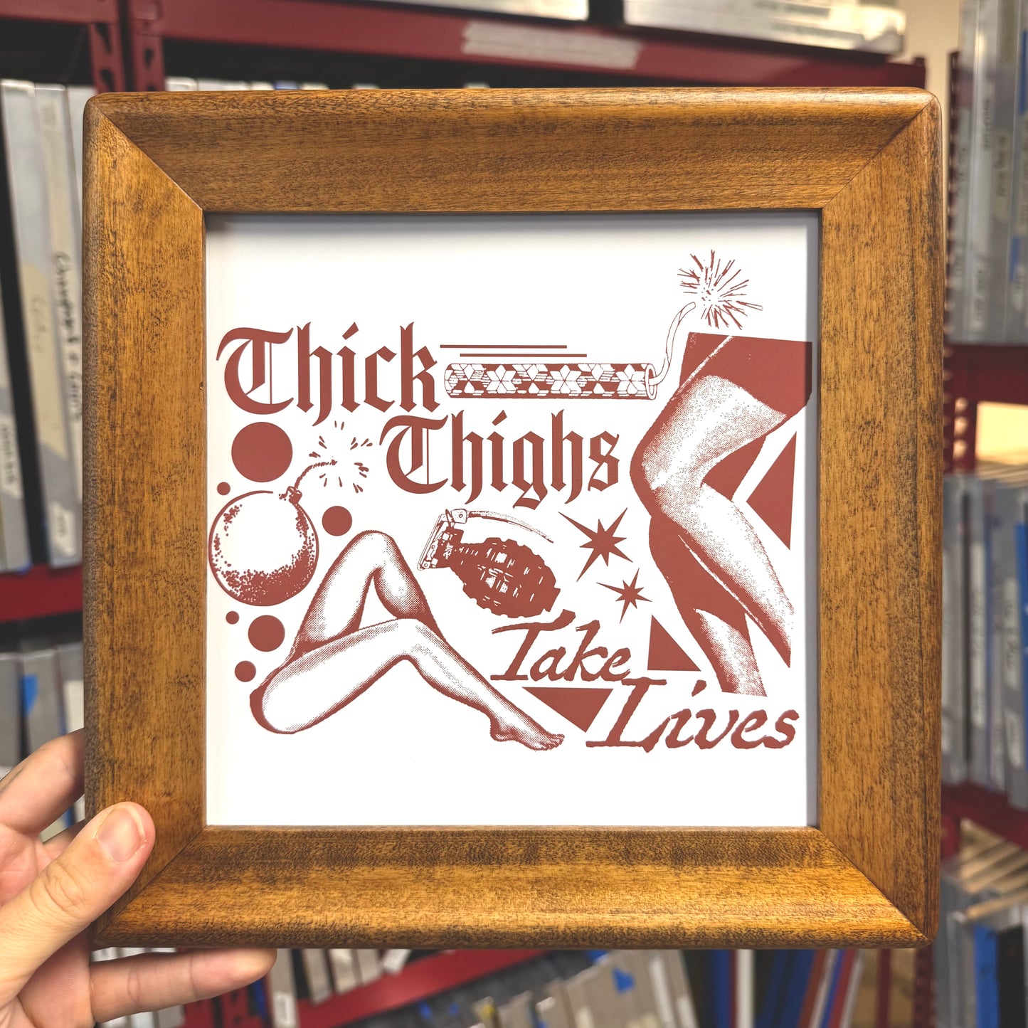 Framed artwork with 'Thick Thighs Take Lives' text and illustrations, held in front of a bookshelf.