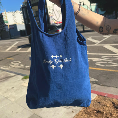 Blue tote bag with branding held by a person on a city street.