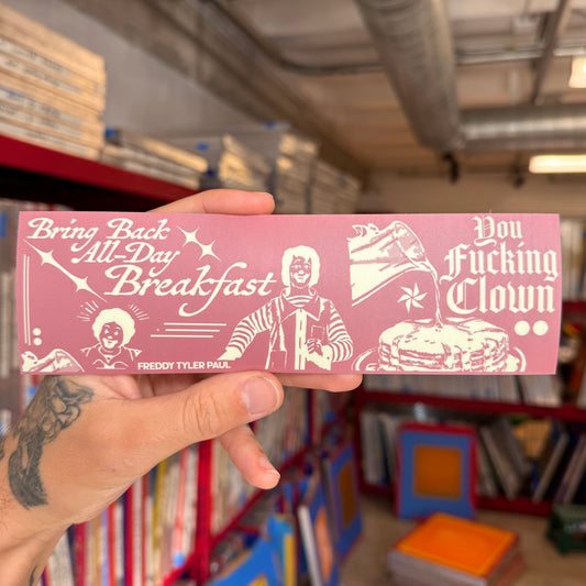 Pink bumper sticker with white text and graphics held in front of a bookshelf.