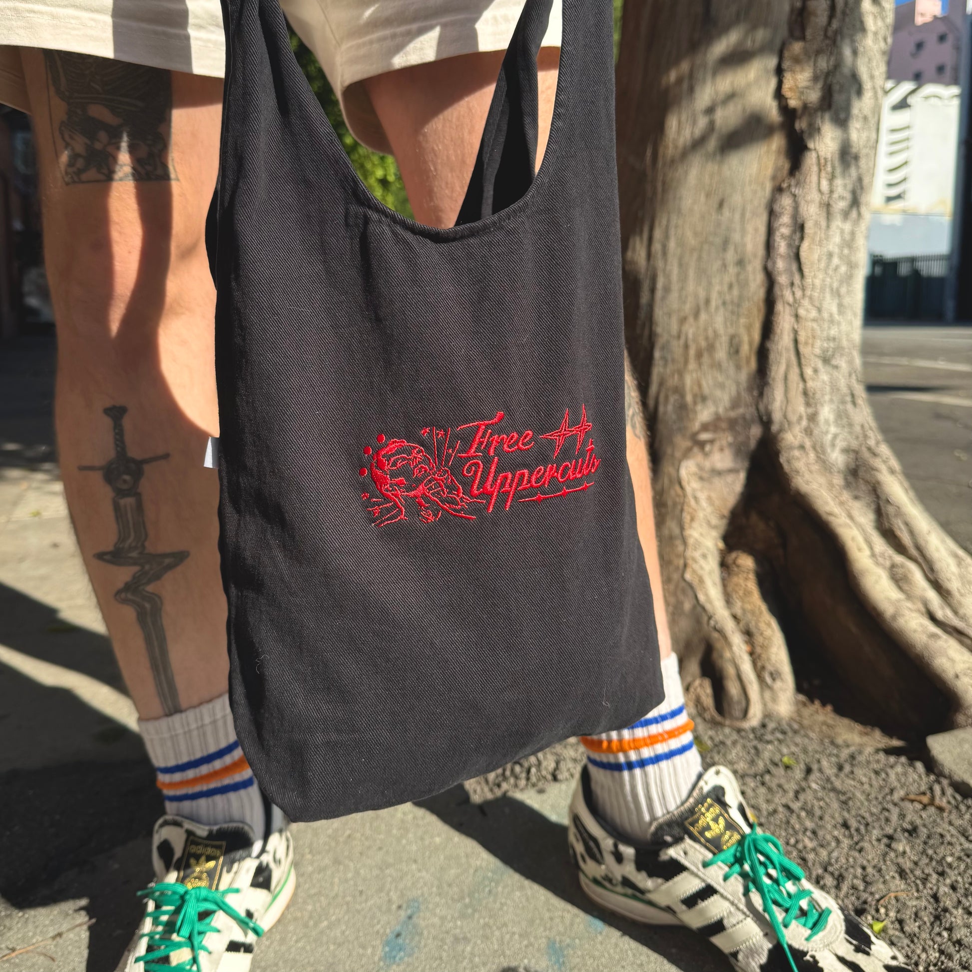 Person holding a black tote bag with red text and graphics, standing next to a tree in an urban setting.