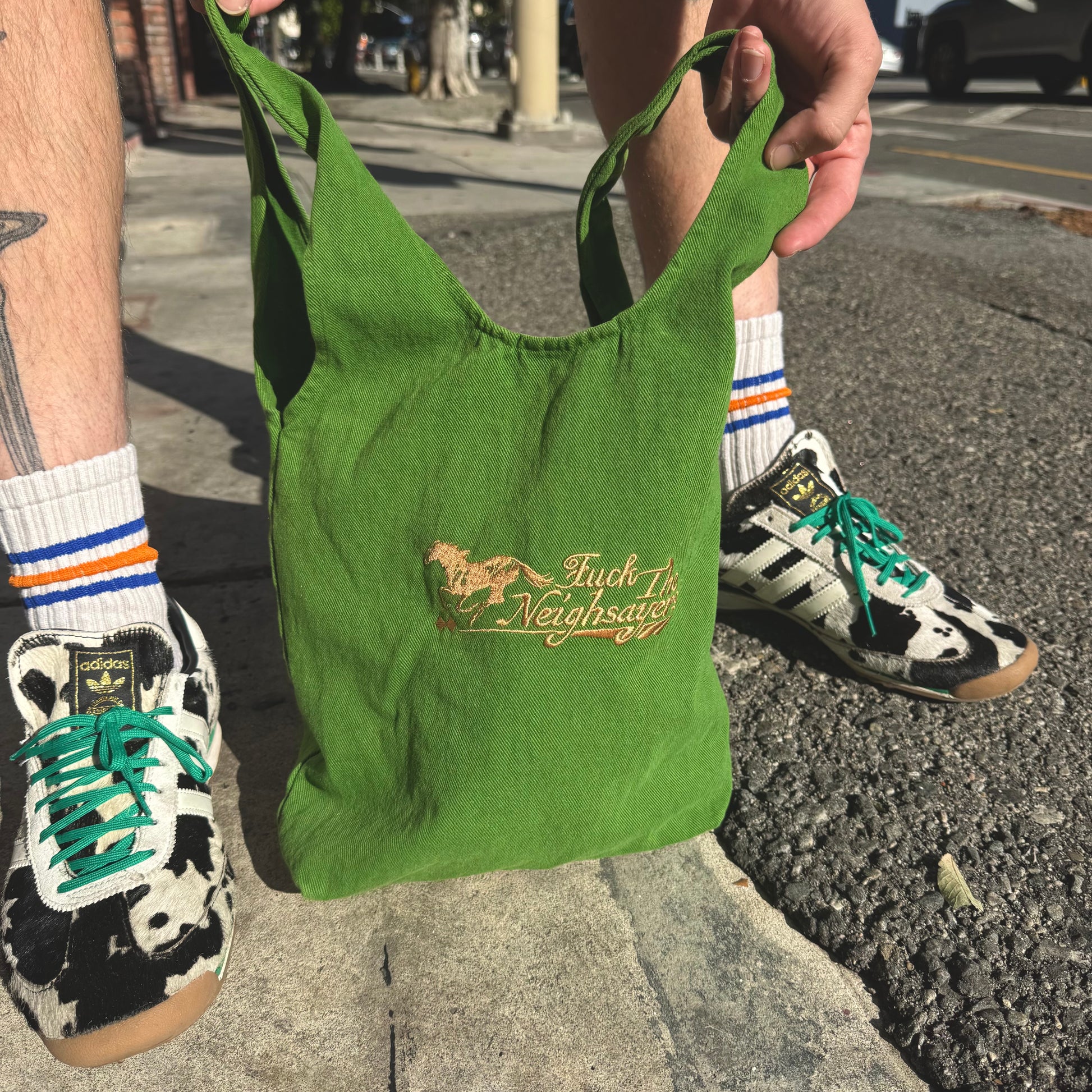 Green bag with text held by a person wearing Adidas shoes and socks on a street.