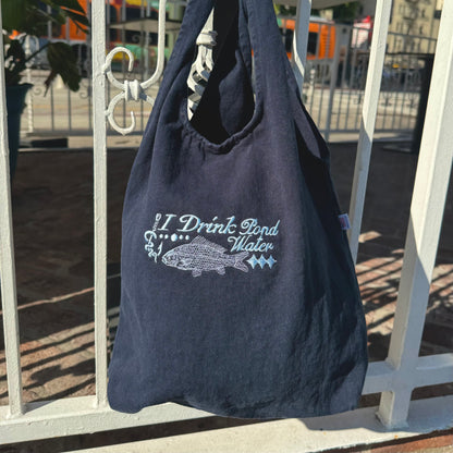 Navy blue tote bag with 'I Drink Pond Water' text and fish graphic on a white metal fence.