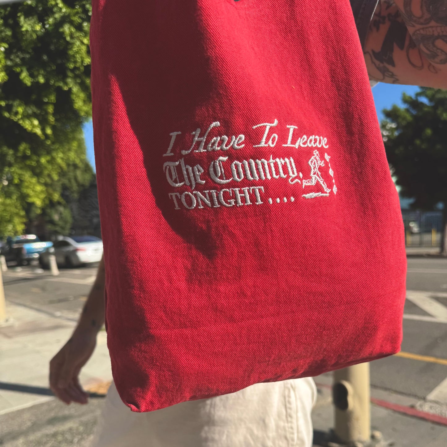 Red bag with white text 'I Have To Leave The Country TONIGHT' held by a person outdoors.
