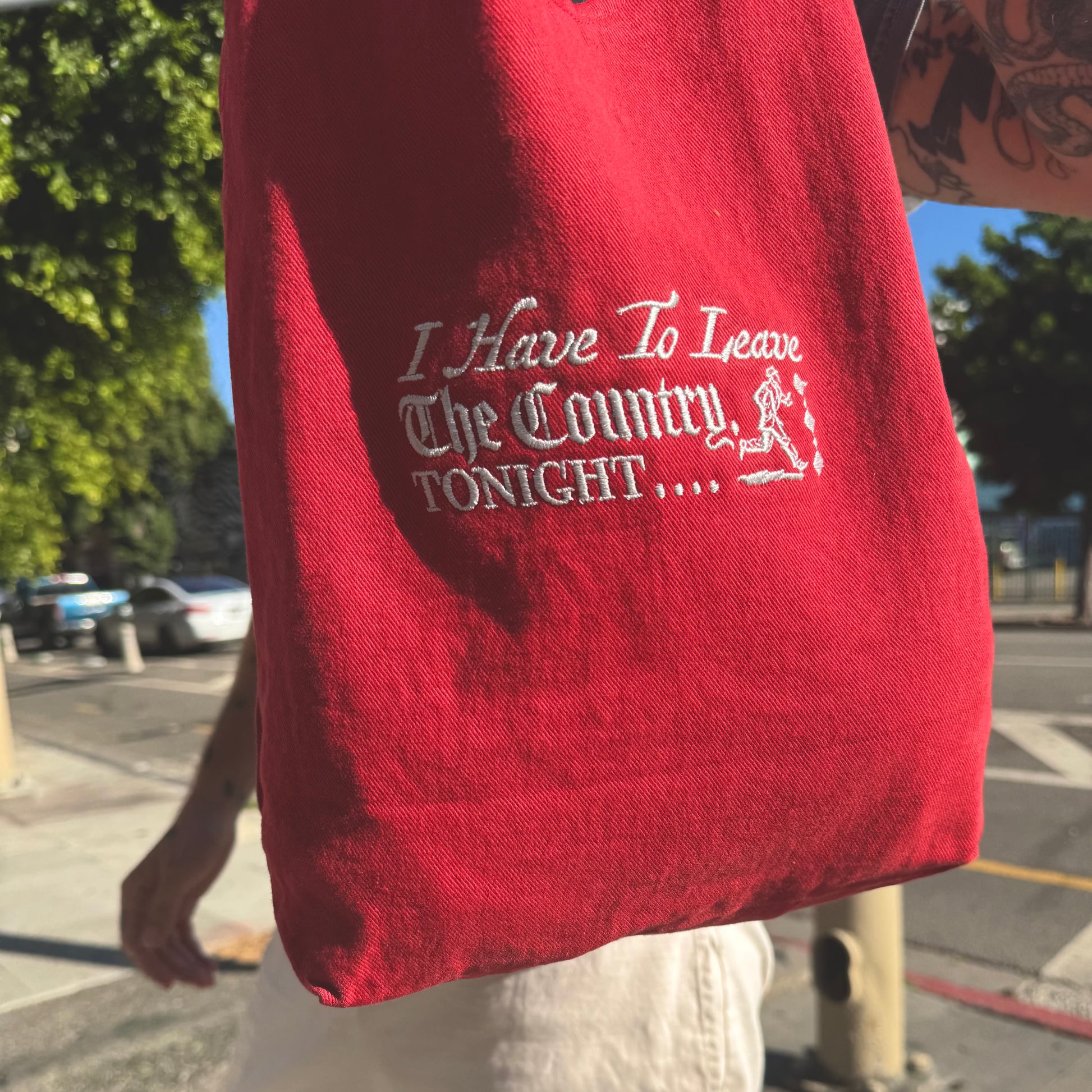 Red bag with white text 'I Have To Leave The Country TONIGHT' held by a person outdoors.