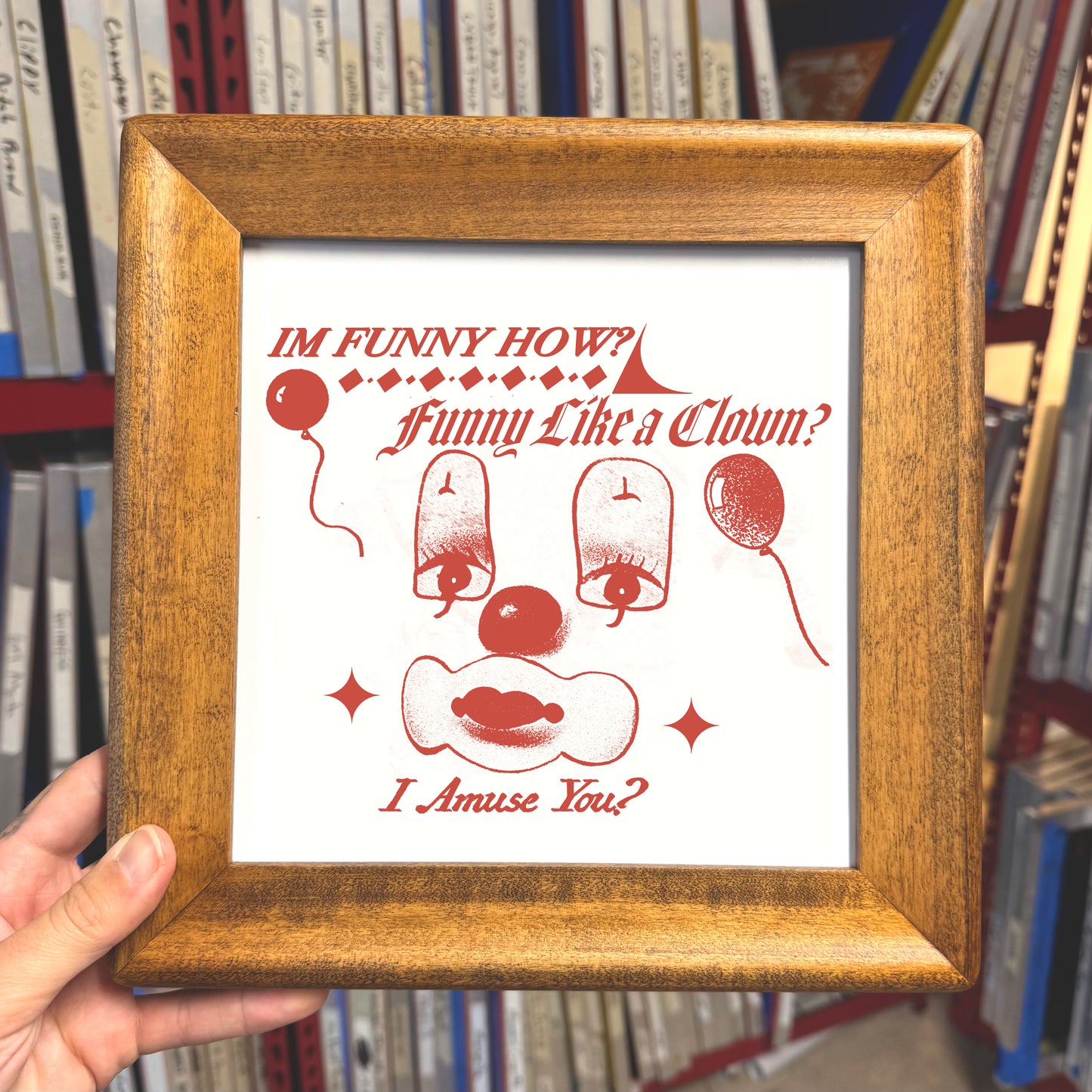 Framed print with humorous text and illustrations held in front of a bookshelf.