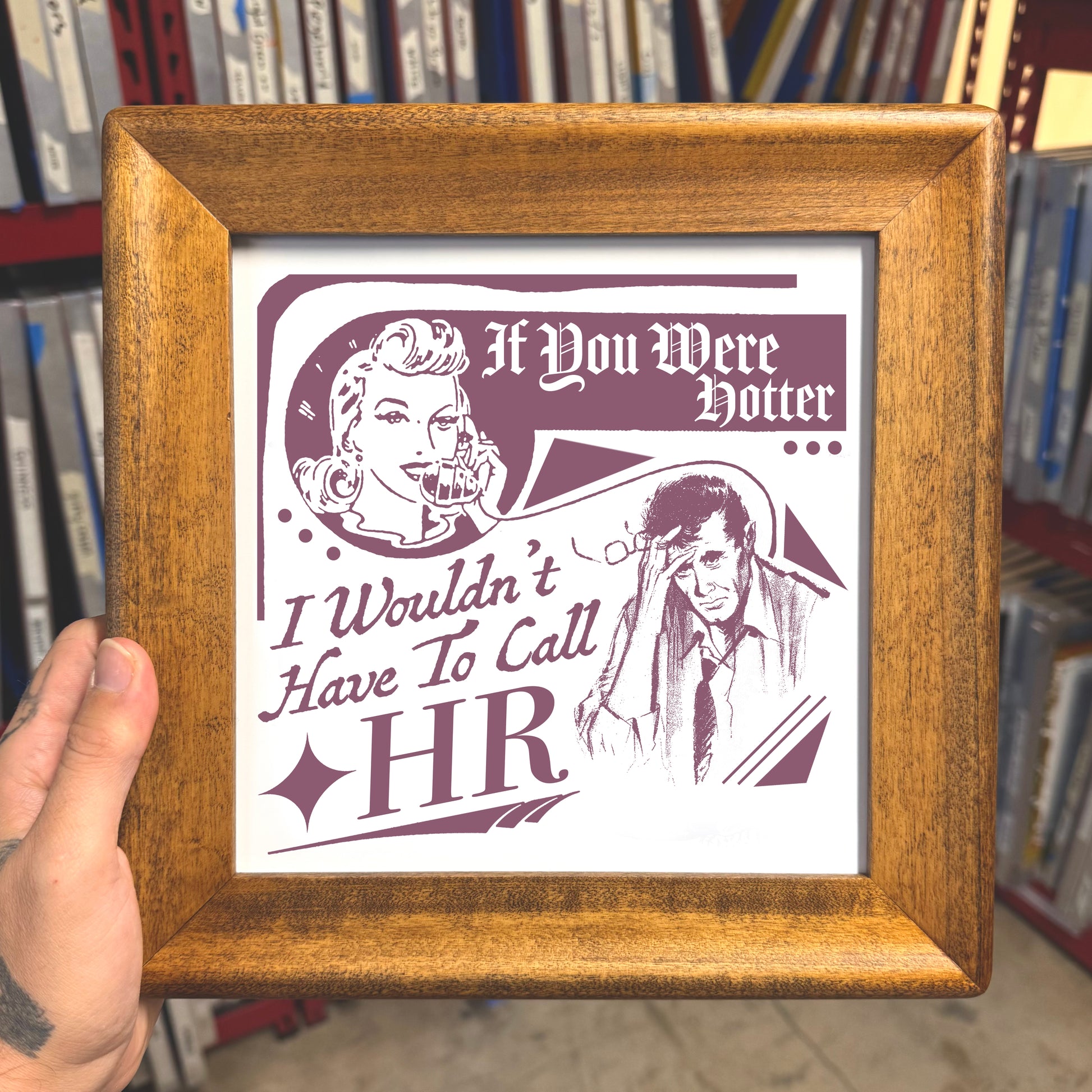 Framed print with humorous illustration and text held in front of a bookshelf.