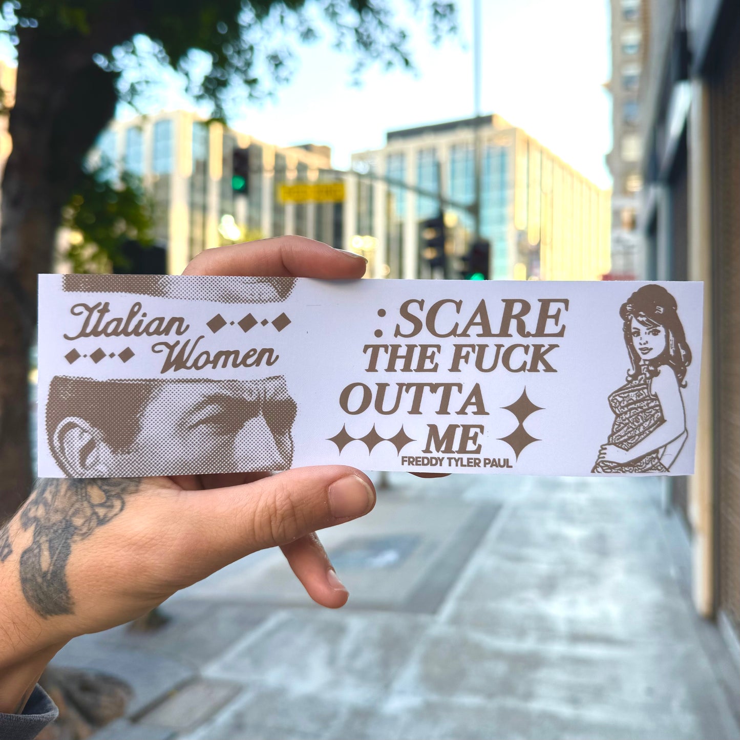 Hand holding a bumper sticker with text and images on a city street background