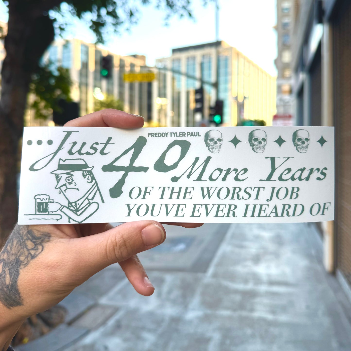 Hand holding a humorous 40 More Years sticker with text and graphics against an urban background.