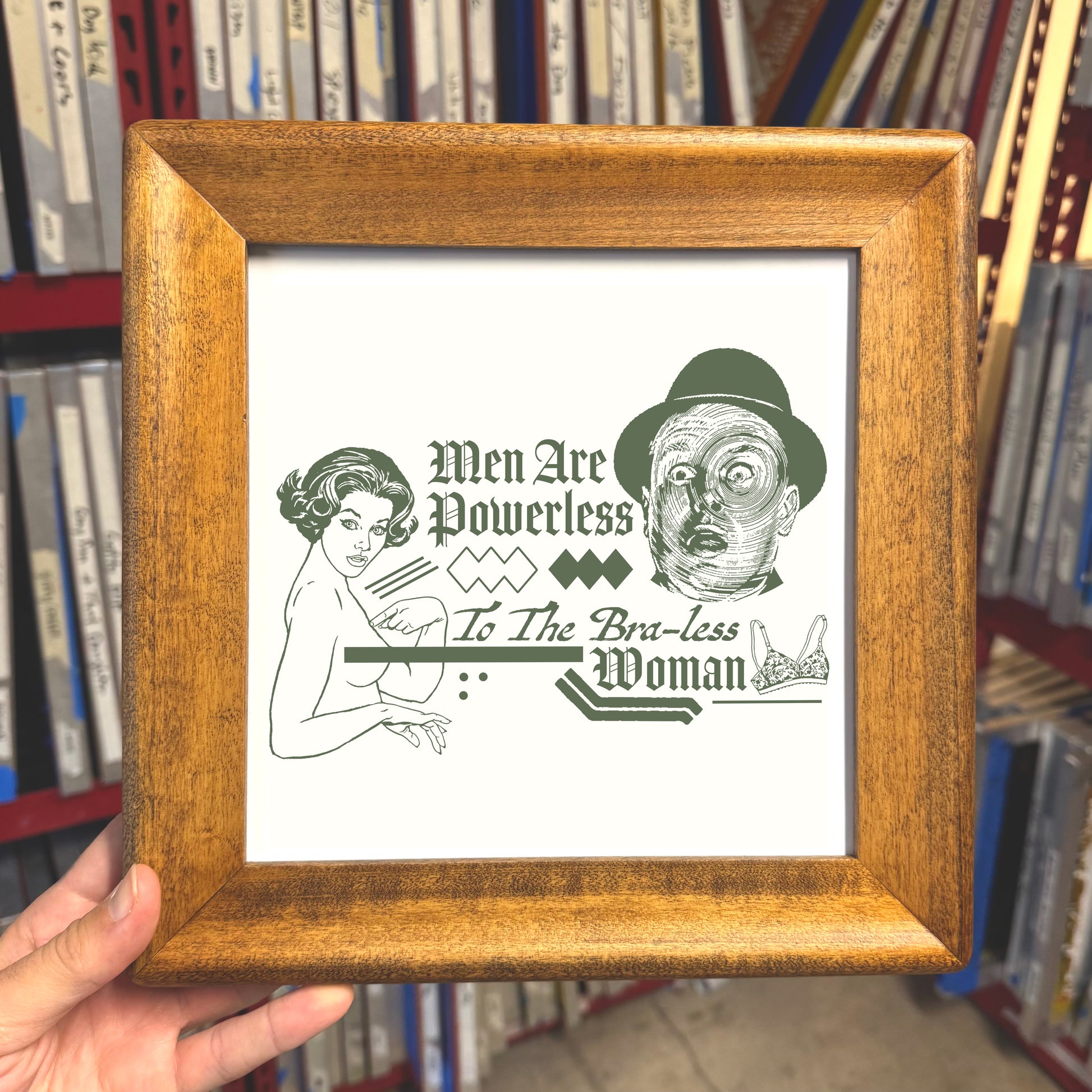 Framed artwork with text and illustrations held in front of a bookshelf.