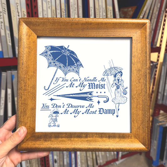 Framed print with blue umbrella design and text held in front of bookshelf