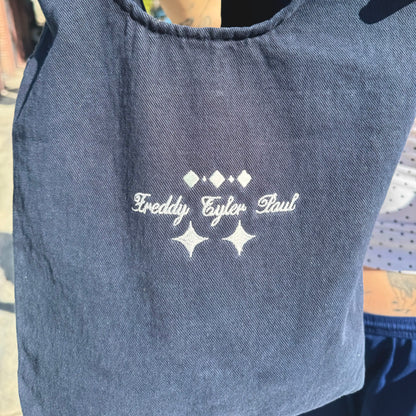 Blue tote bag with embroidered text and star designs on a person wearing it.
