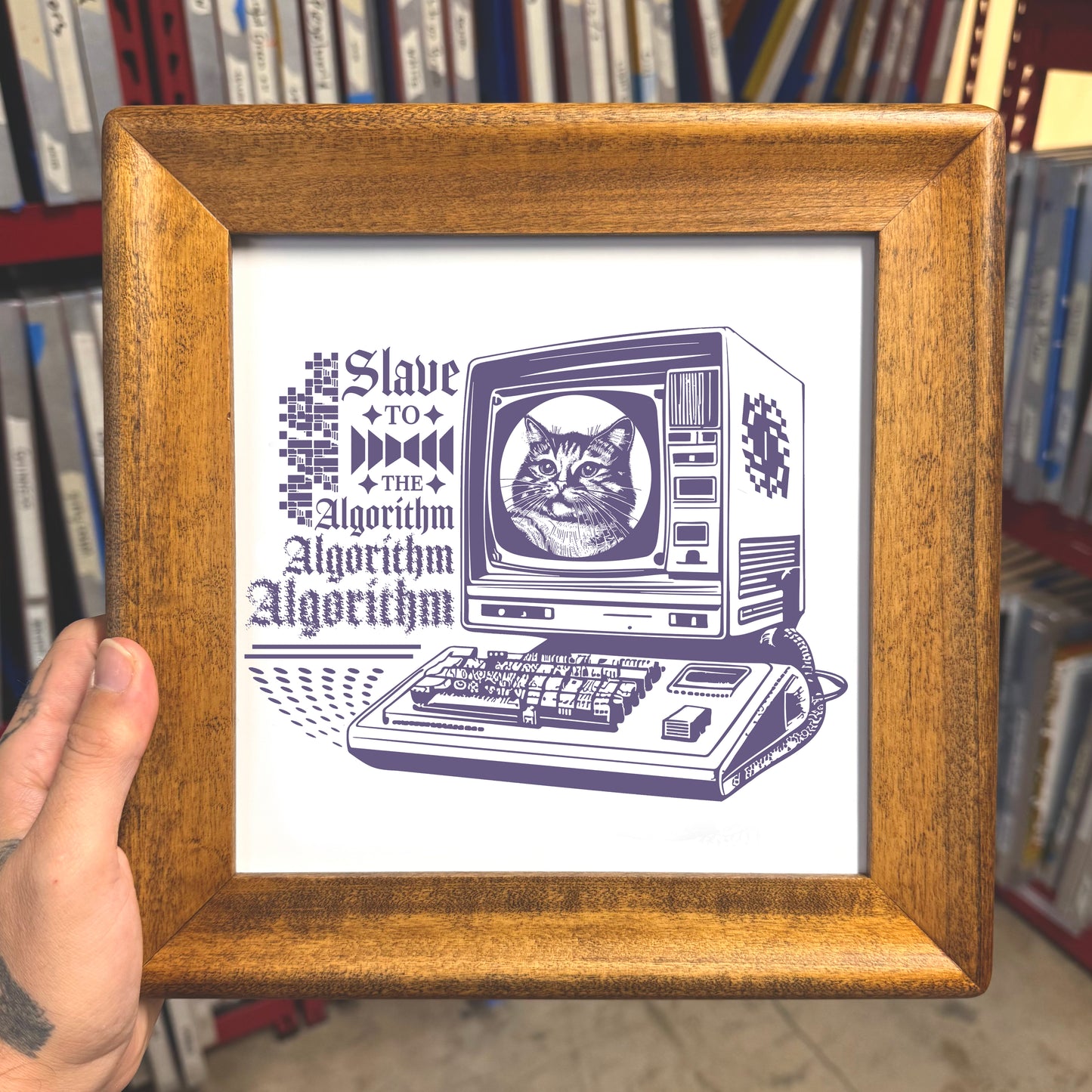 Framed artwork with a cat on a computer monitor and text 'Slave to the Algorithm' held in front of bookshelves.