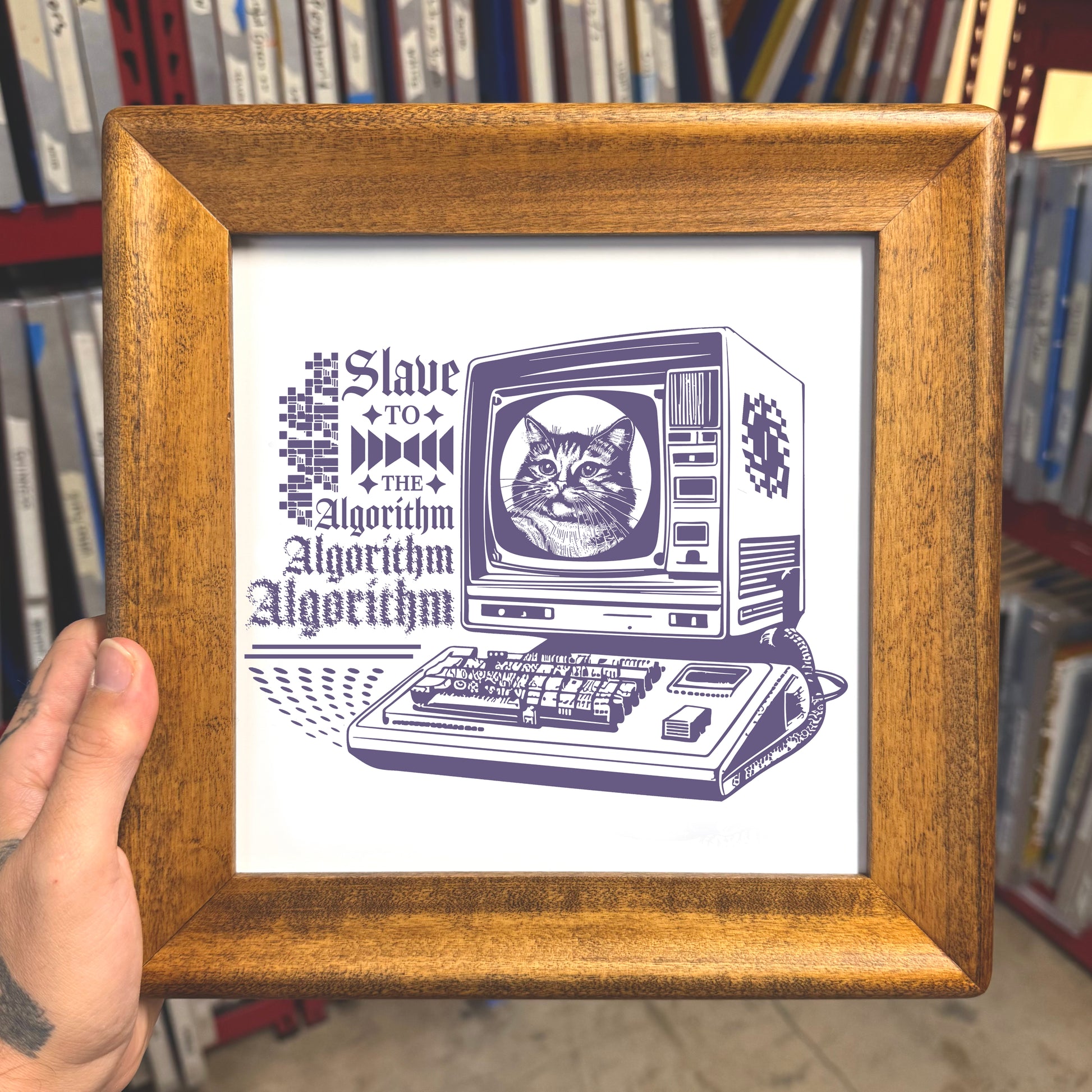 Framed artwork with a cat on a computer monitor and text 'Slave to the Algorithm' held in front of bookshelves.