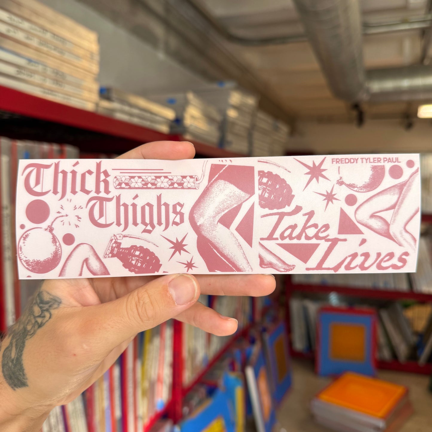 Hand holding a pink and white sticker with text and graphics in a bookstore setting