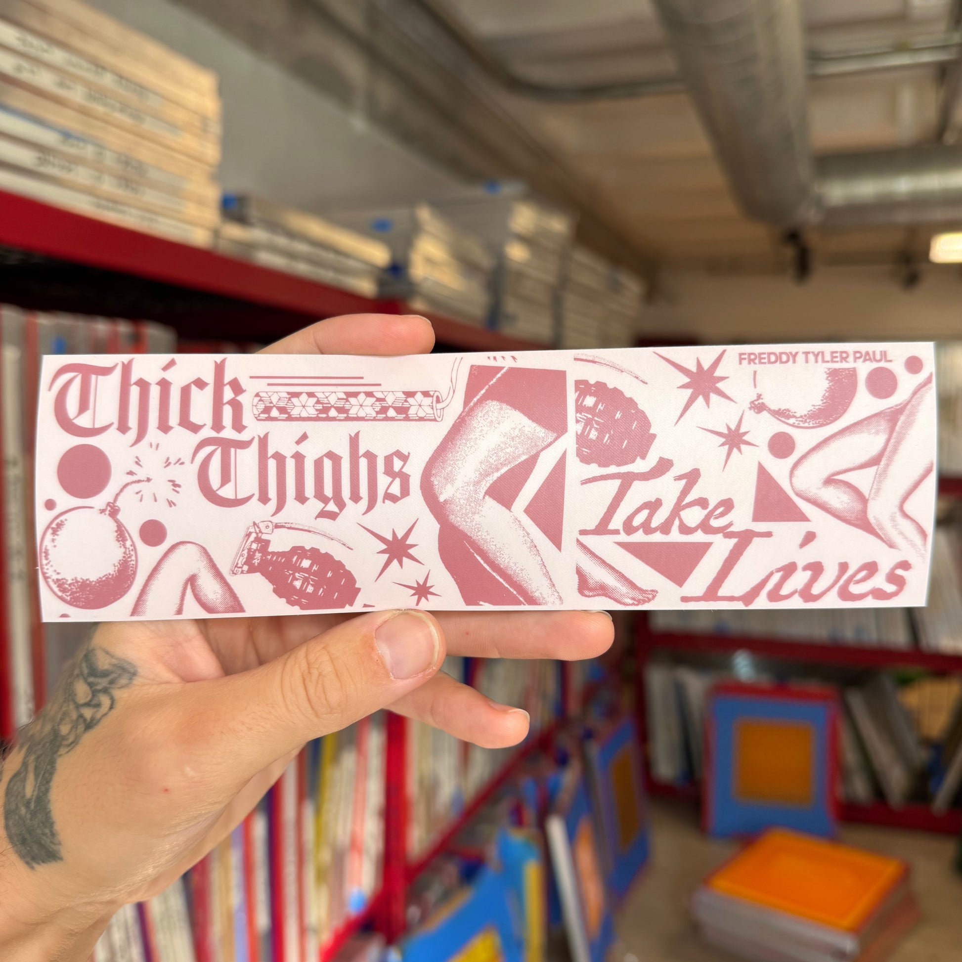 Hand holding a pink and white sticker with text and graphics in a bookstore setting