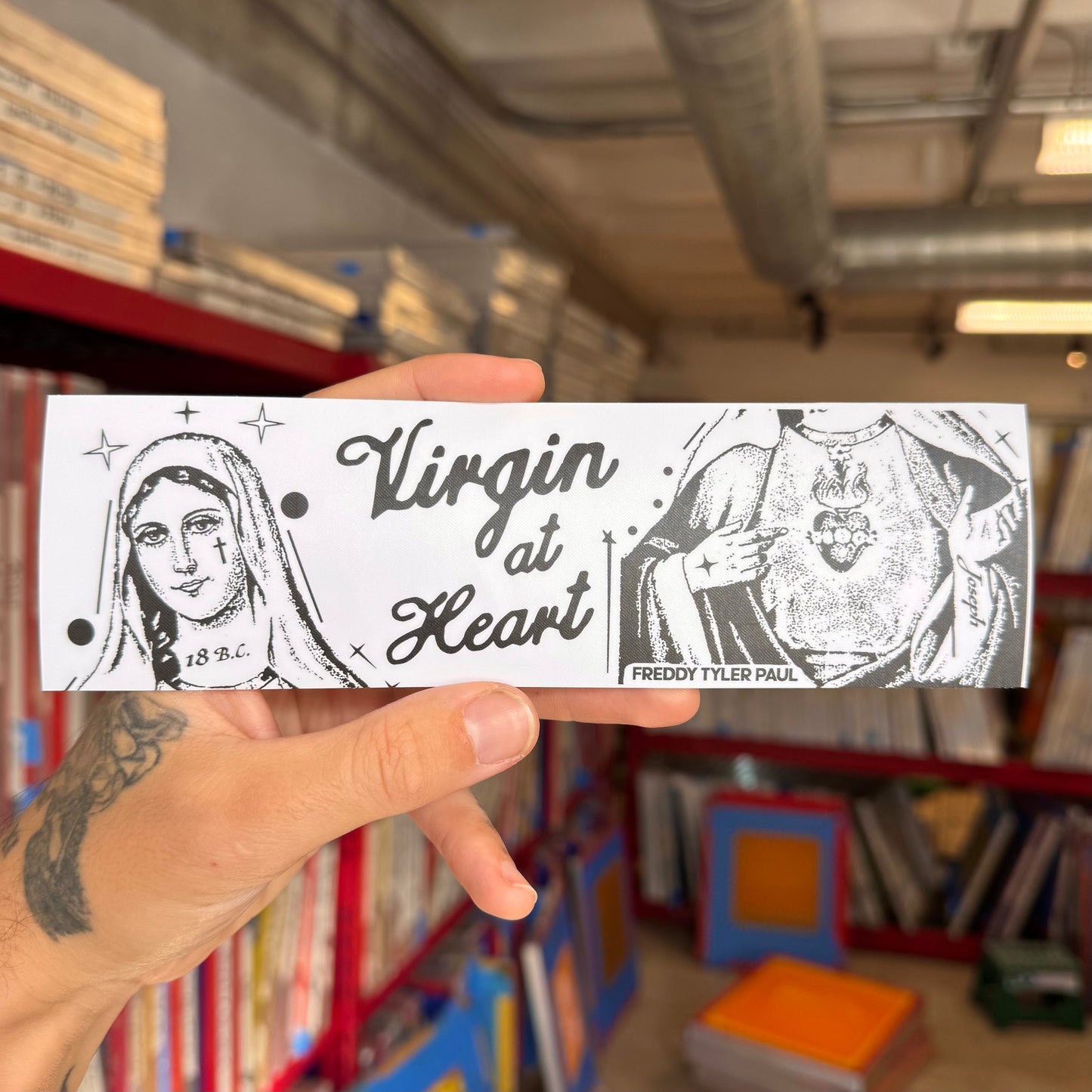 Hand holding a 'Virgin at Heart' bumper sticker in a bookstore setting