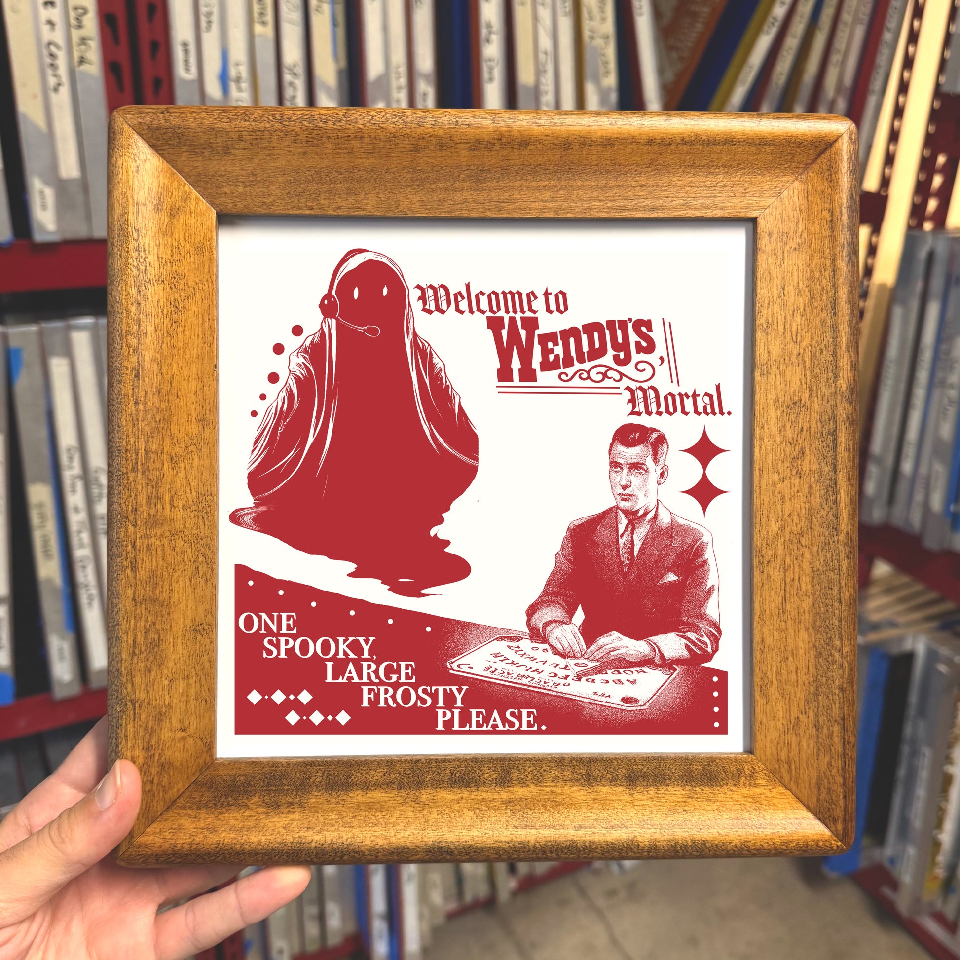 Framed print with red illustration of a ghost and man, text 'Welcome to Wendy's Mortal', against a bookshelf background.