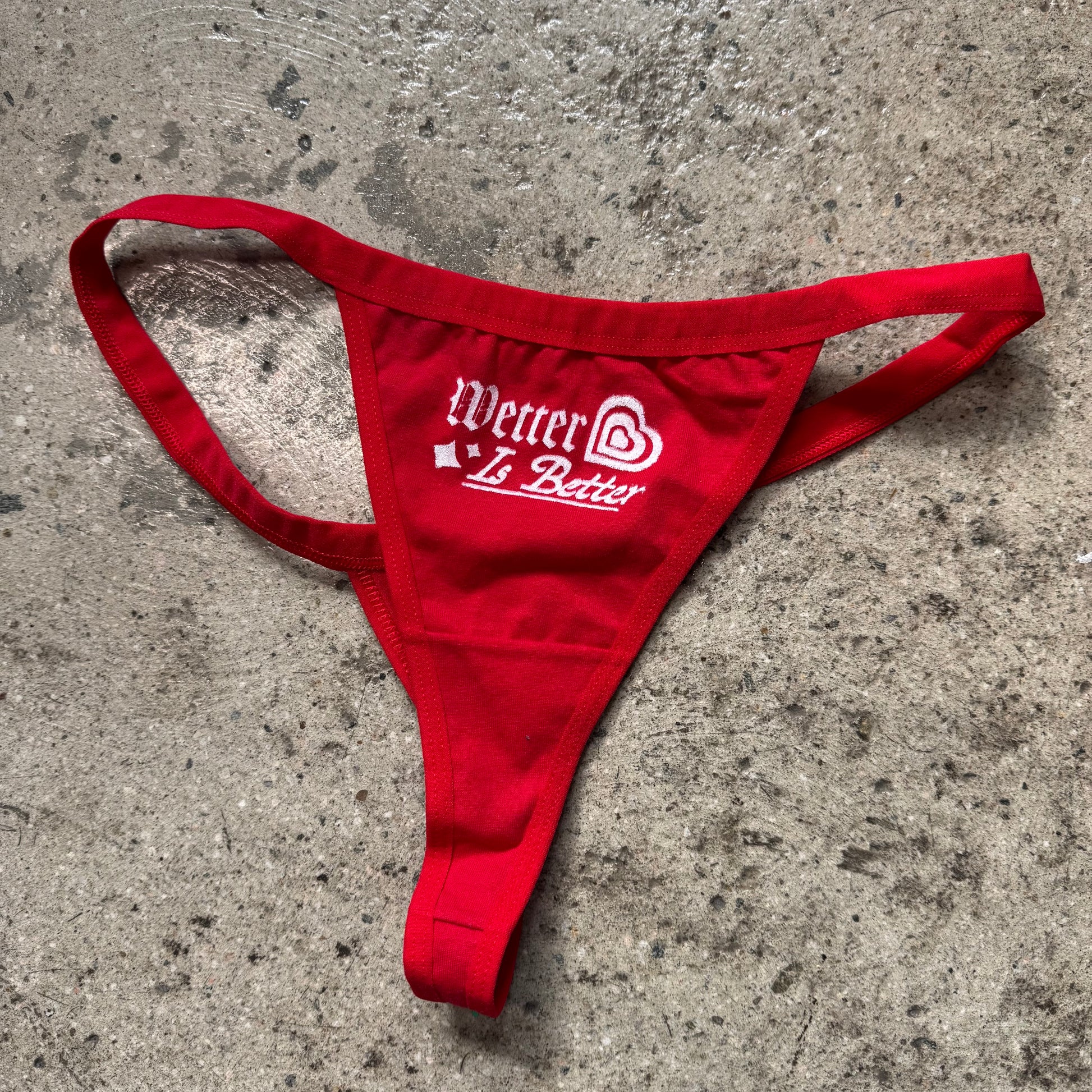 Red thong with 'Wetter is Better' text on a concrete surface