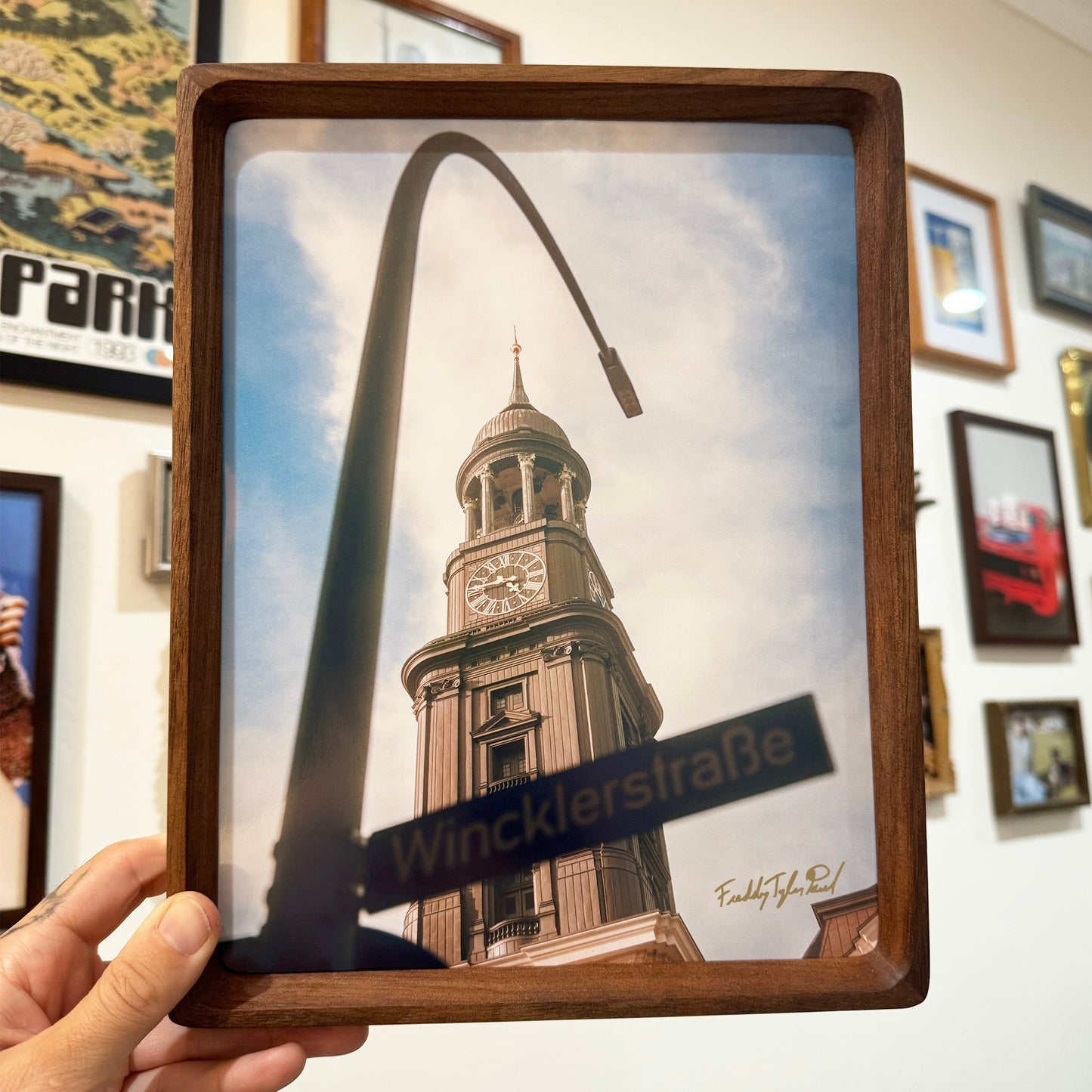 Framed picture of a clock tower with 'Wincklerstraèe' street sign held by a hand against a wall with other framed pictures.