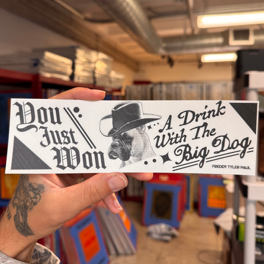 Hand holding a bumper sticker with text and a dog illustration in an indoor setting.