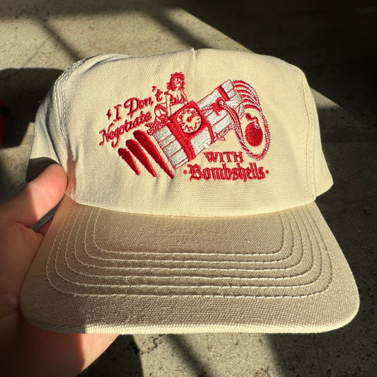 Beige cap with red embroidered text and design held by a hand on a concrete surface.