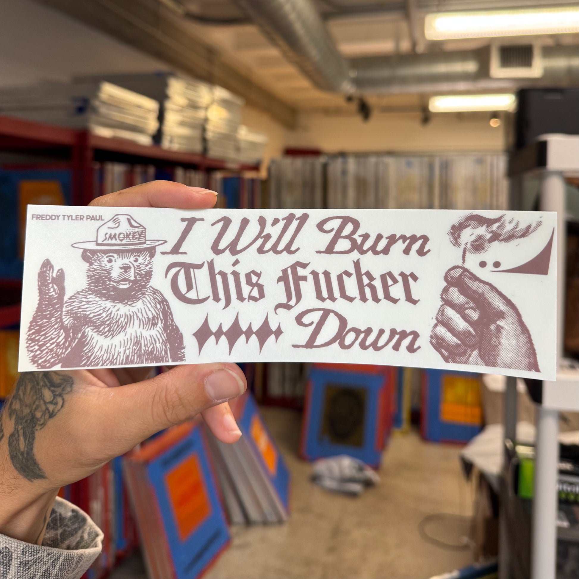 Hand holding a sticker with graphic text and bear illustration in an indoor setting.