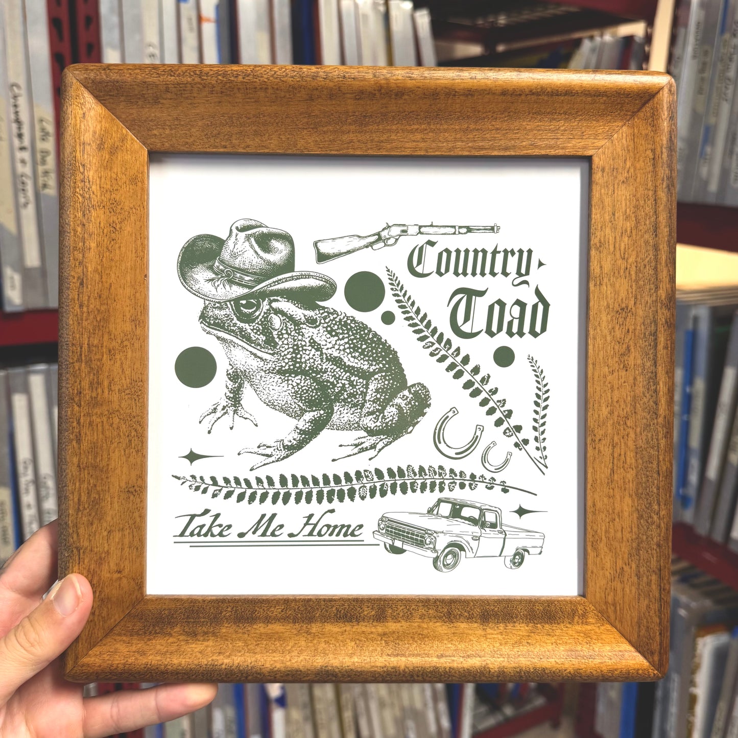 Framed print with a frog wearing a cowboy hat and text 'Country Toad Take Me Home' held in front of bookshelves.