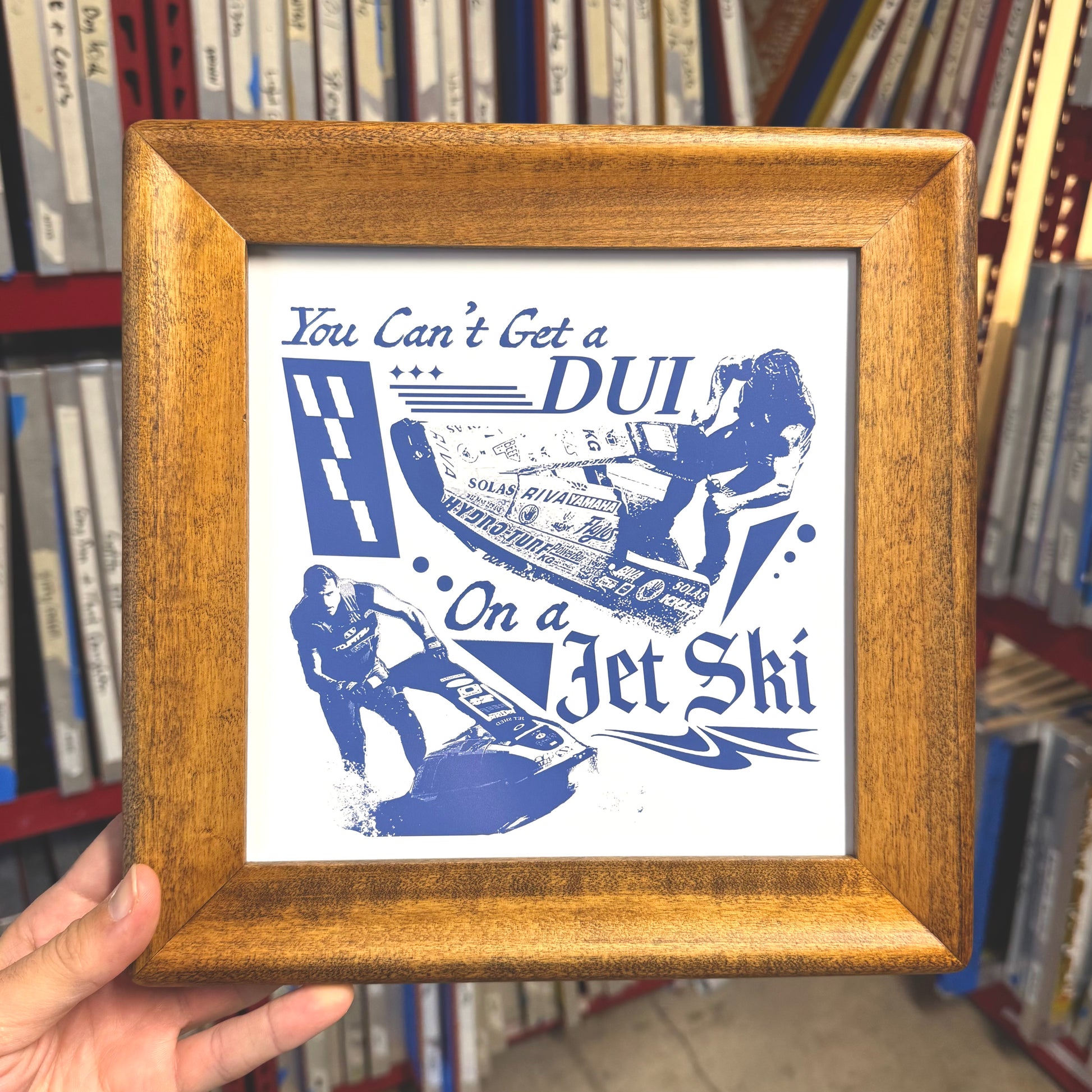 Framed poster with blue text and graphics on a white background, held in front of a bookshelf.