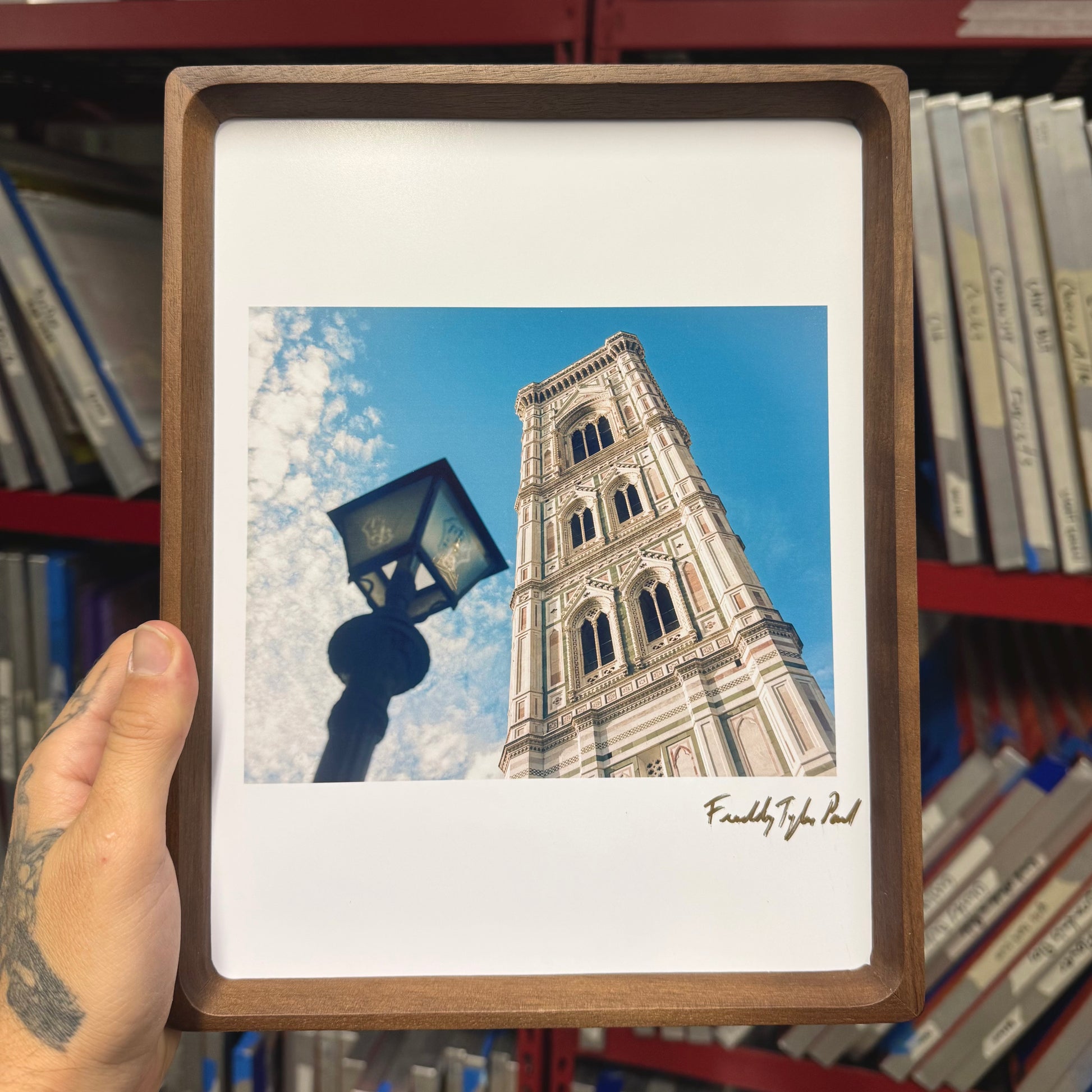 Framed picture of a tall tower with a blue sky, held in front of a bookshelf.