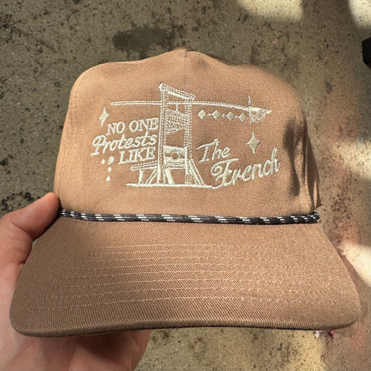 Brown cap with white embroidered text held against a concrete background