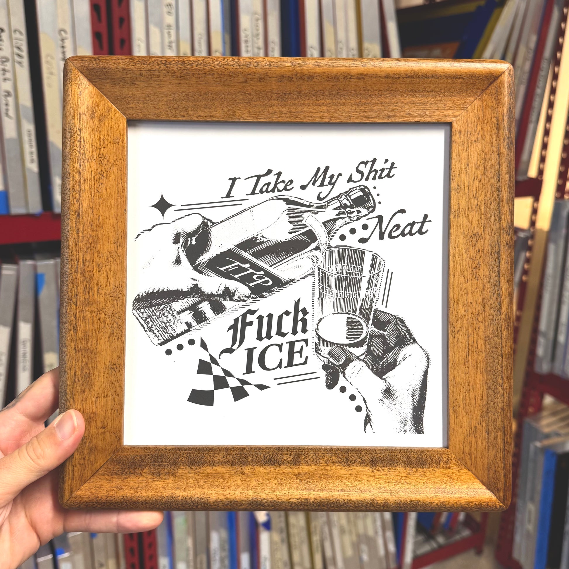 Framed artwork with black and white illustration and text held in front of a bookshelf.