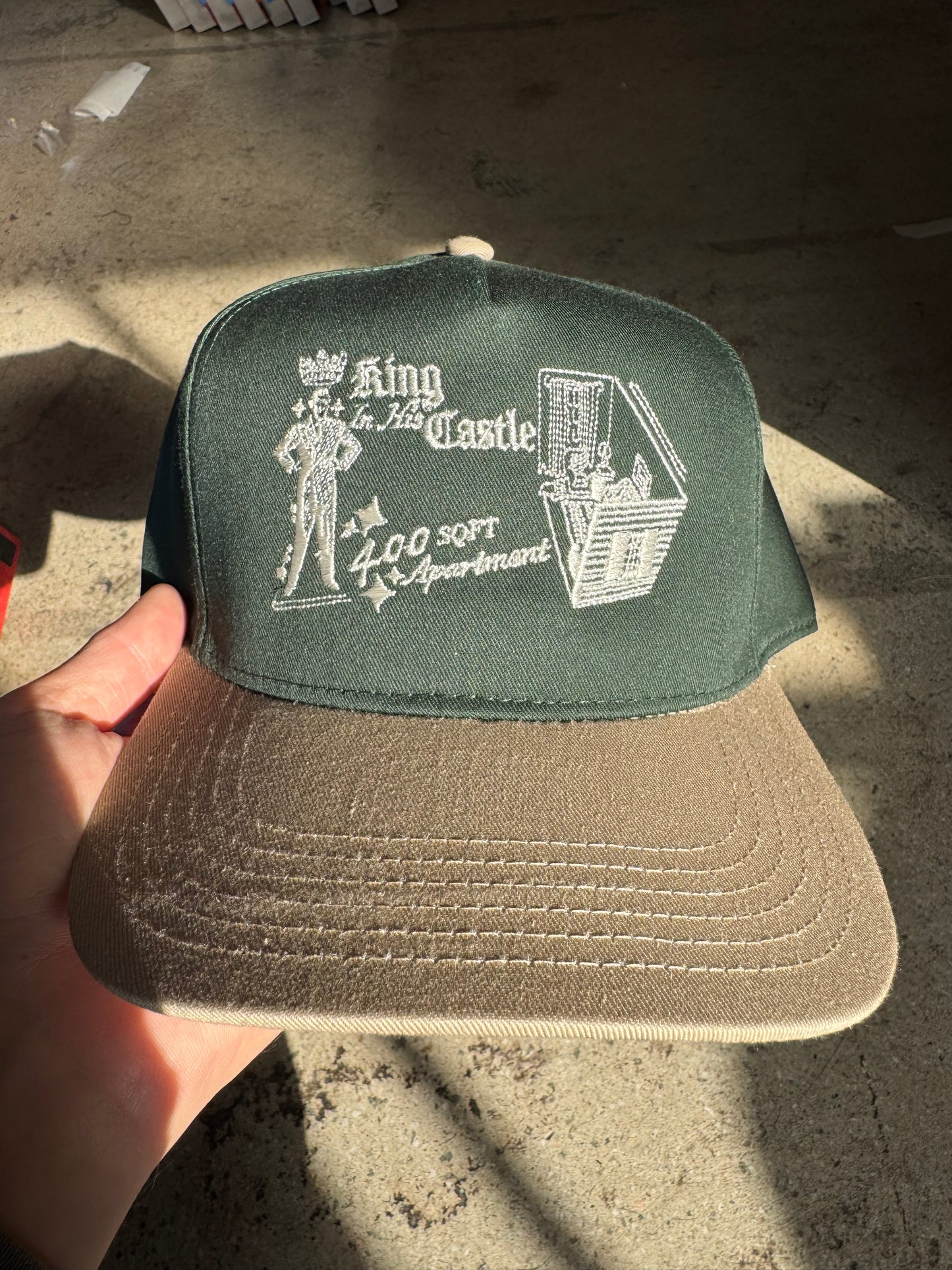 Beige cap with green patch featuring text and graphics held by a hand on a concrete surface.