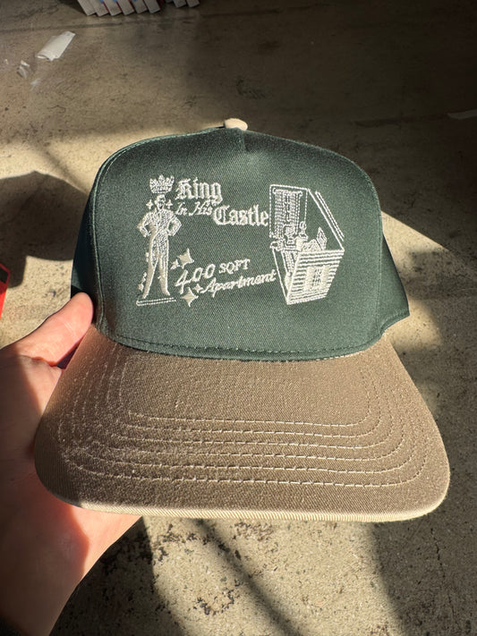 Beige cap with green patch featuring text and graphics held by a hand on a concrete surface.