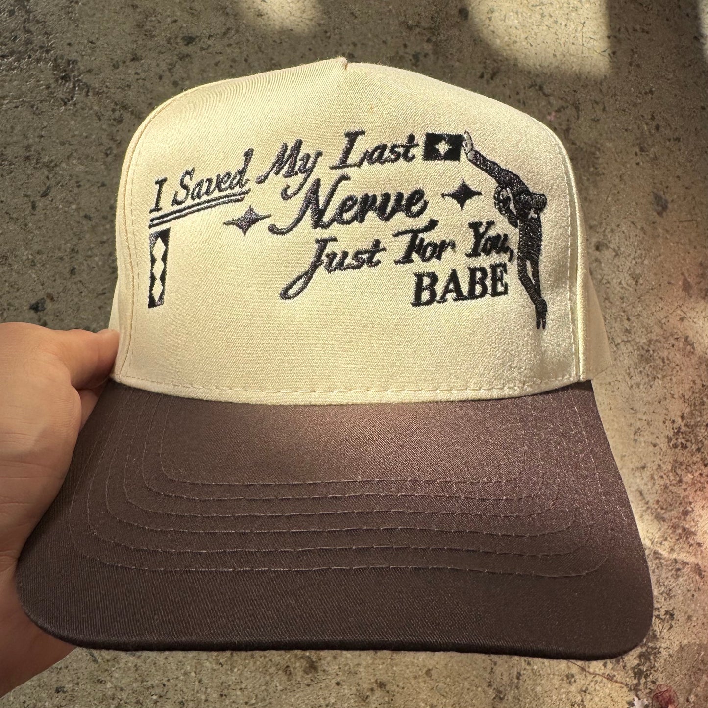 Cap with text 'I Saved My Last Nerve Just For You, Babe' held by a hand on a textured surface.