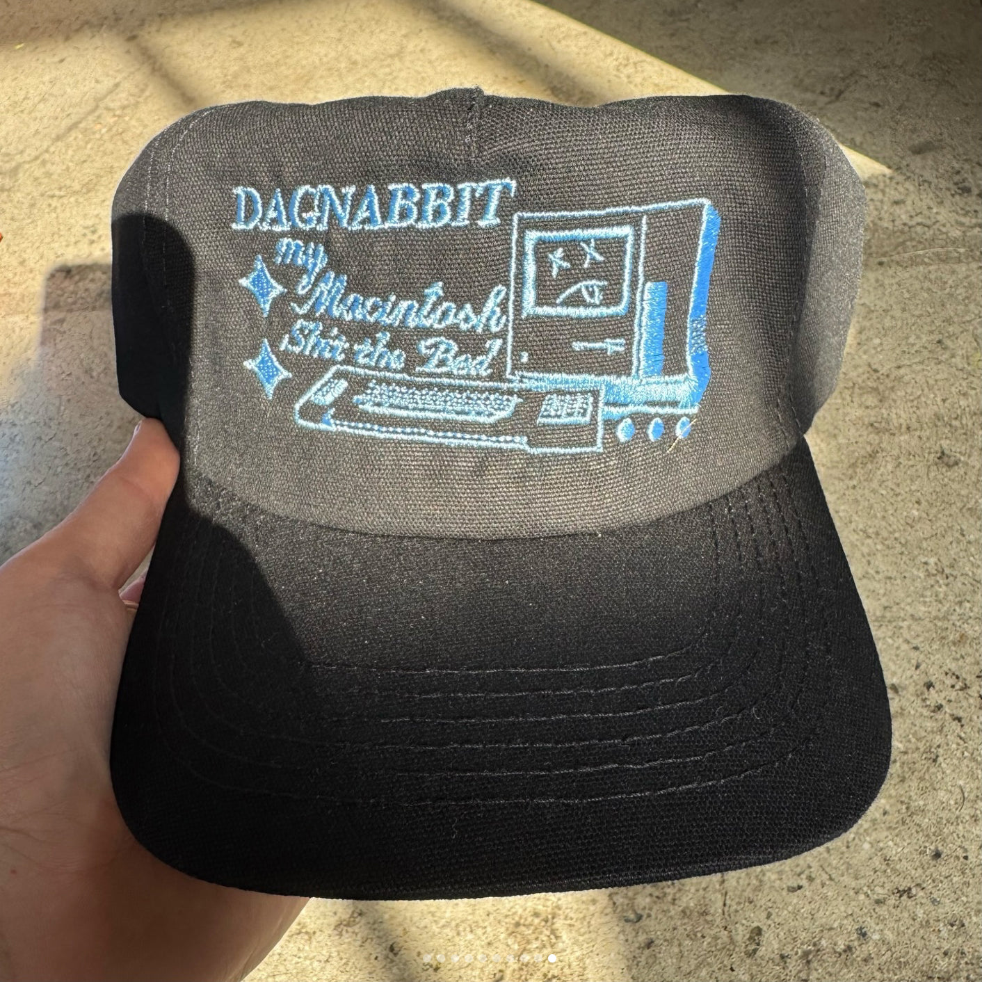 Black cap with blue text and graphics held by a hand on a concrete surface