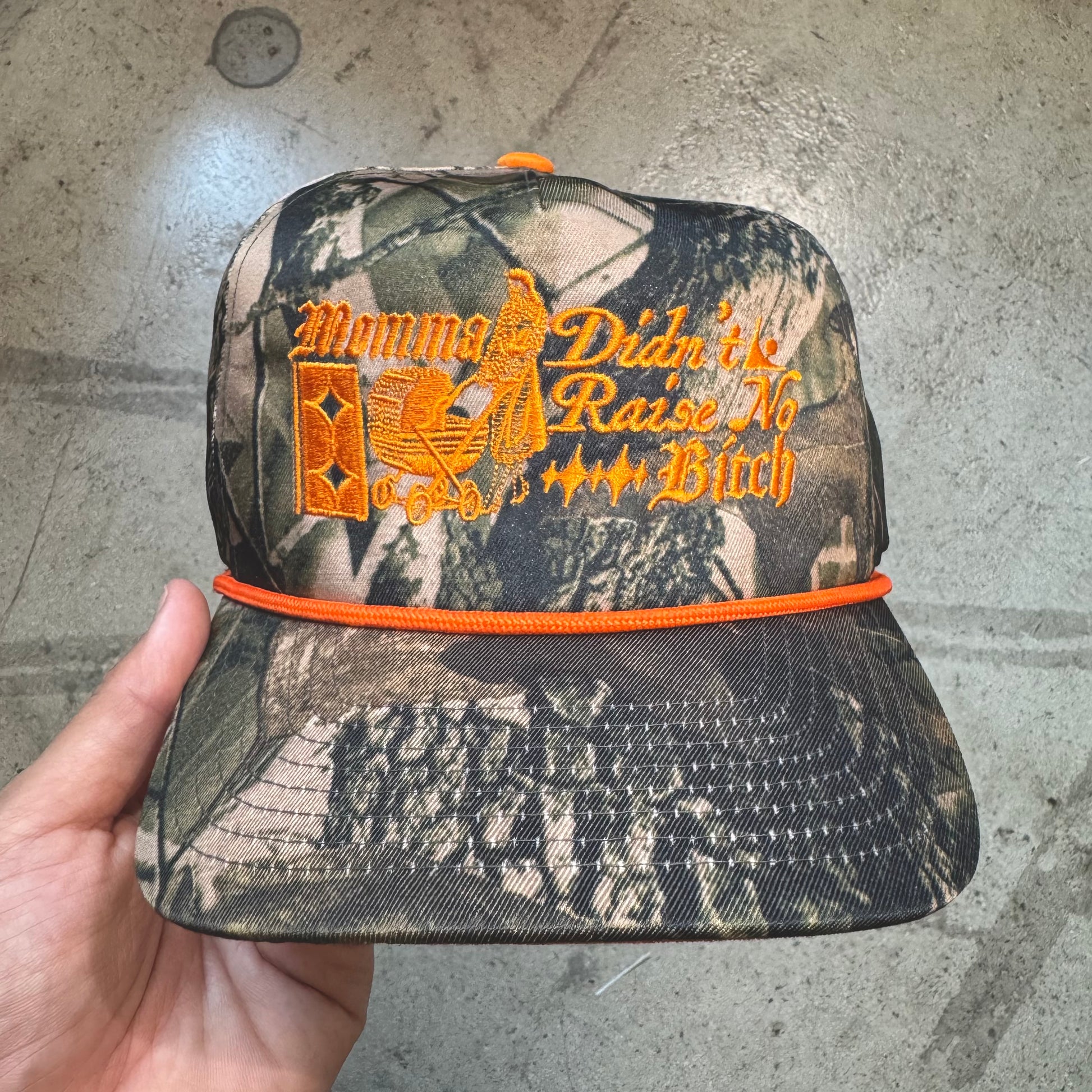 Camouflage cap with orange text held by a hand on a concrete floor.