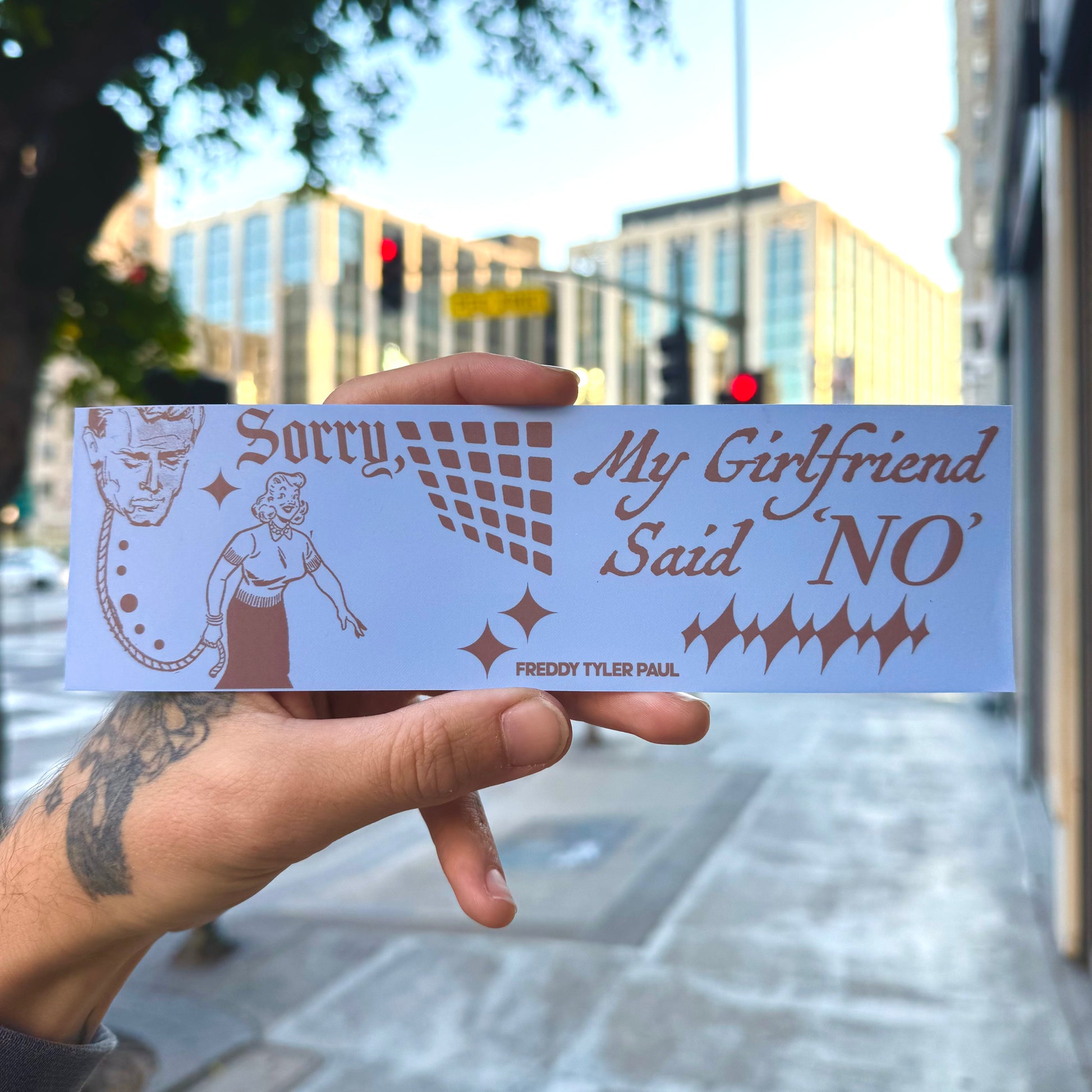 Hand holding a bumper sticker with text and graphics outdoors.