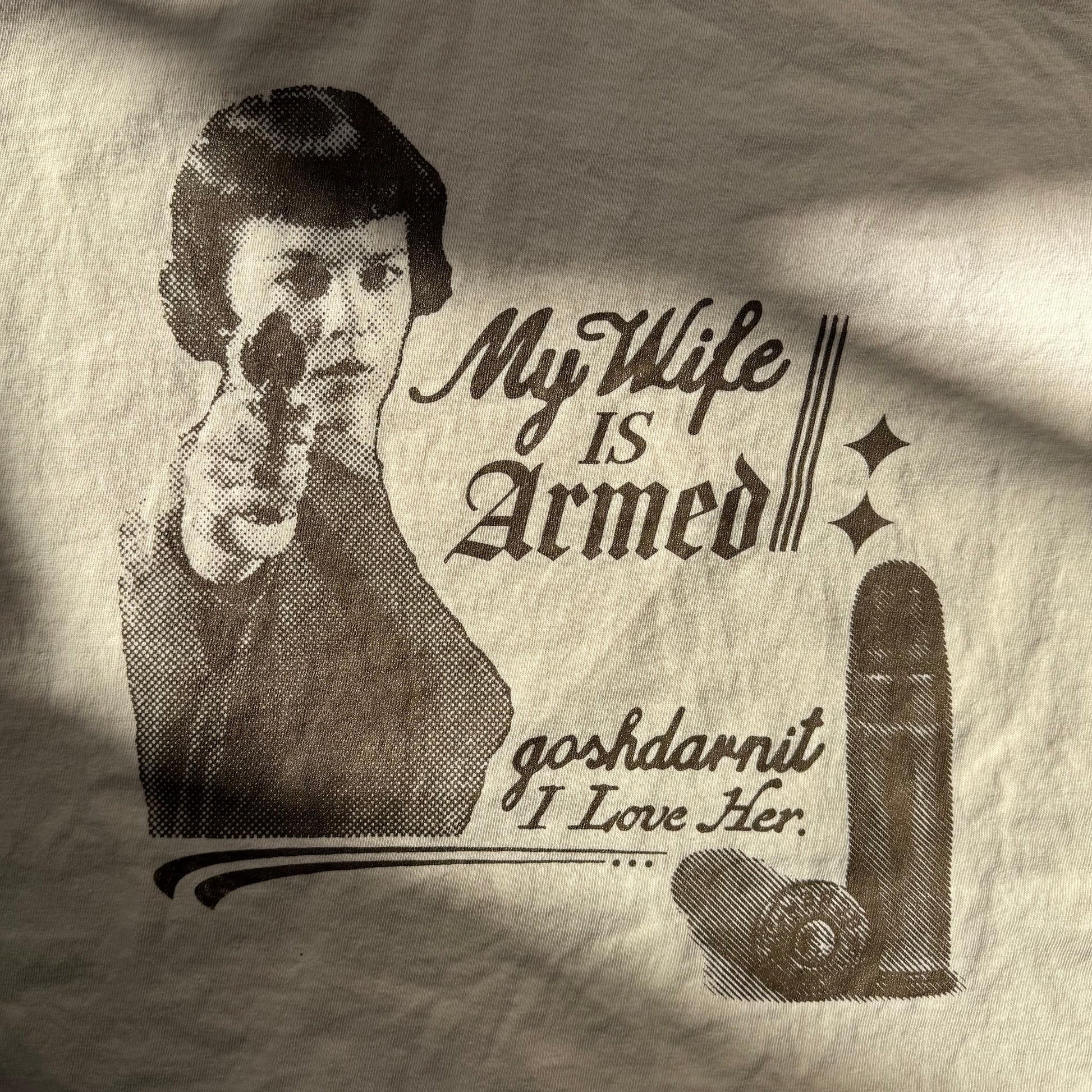 T-shirt with graphic of a woman holding a gun and text 'My Wife is Armed goshdarnit I Love Her' on a beige background.