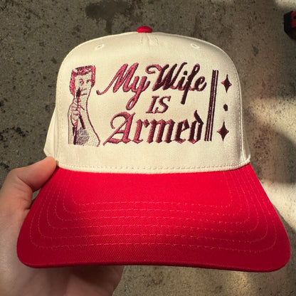 Red and white cap with 'My Wife is Armed' text and graphic held by a hand against a textured background.