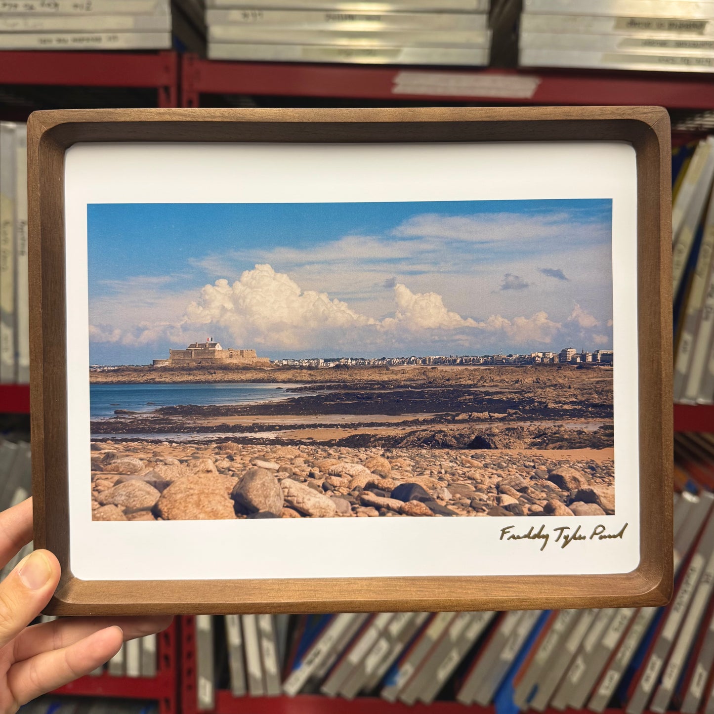 Framed photograph of a coastal scene with a castle-like structure, held in front of a stack of books.