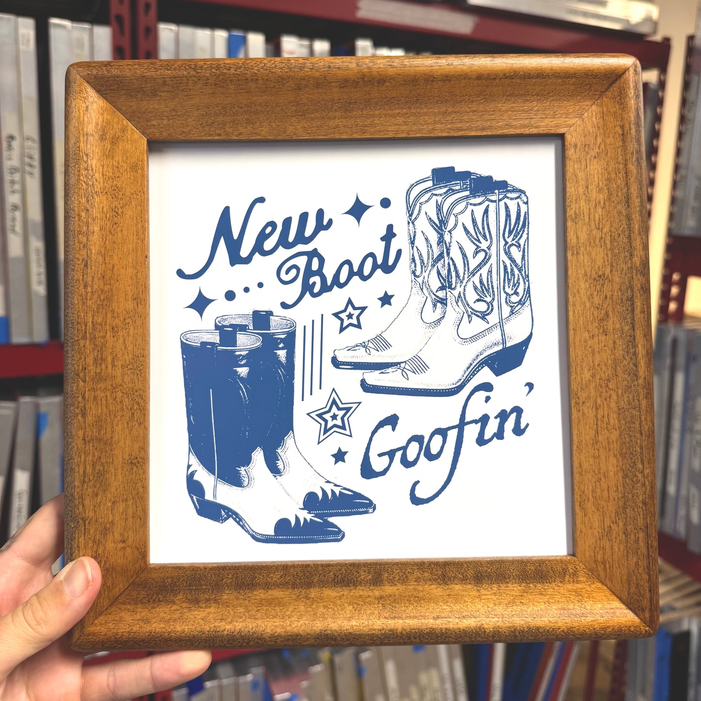 Framed print with blue cowboy boots and text 'New Boot Goofin'' held in front of a bookshelf.