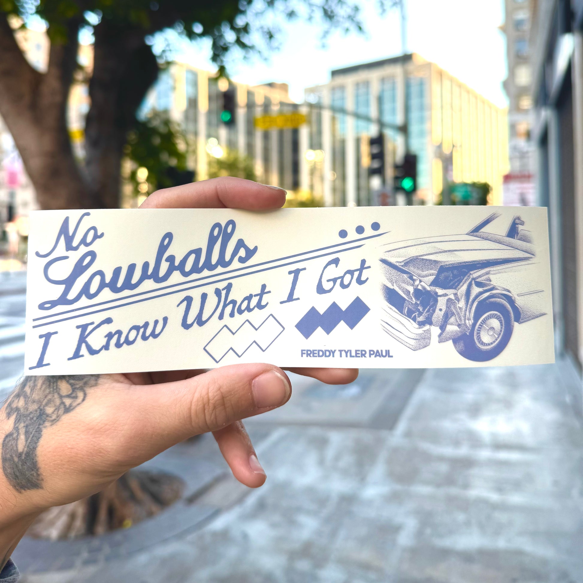 Hand holding a bumper sticker with text and a car illustration outdoors.